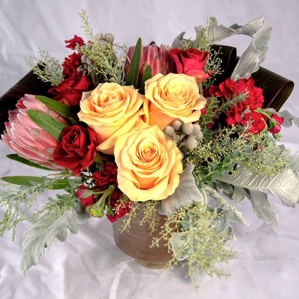 Celine Celine - Amber-yellow roses complimented with darker hues of red protea flowers, magenta stocks and greenish-gray eucalyptus. Low compact design set in ceramic vase. (Approx. size 10"w x 12"h x 10"d)
