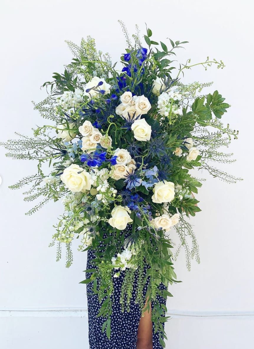 Standing Spray - This oblong hanging floral spray adds a touch of elegance to celebrations of life, services, and even weddings. Designed in a graceful white and green color scheme, it features a lush blend of roses, spray roses, stock, and fresh greenery, creating a timeless and sophisticated look. Suspended from a 4.5ft stand, this arrangement brings height and beauty to any setting. Pictured in its deluxe version, it’s perfect for those looking to make a lasting impression with classic, refined floral styling.   Note: Standard Funeral Spray will be made with white flowers and greenery. Please specify in the notes section if there is another color that you would like added in addition to the white flowers and greenery. Pictured is the Deluxe. 