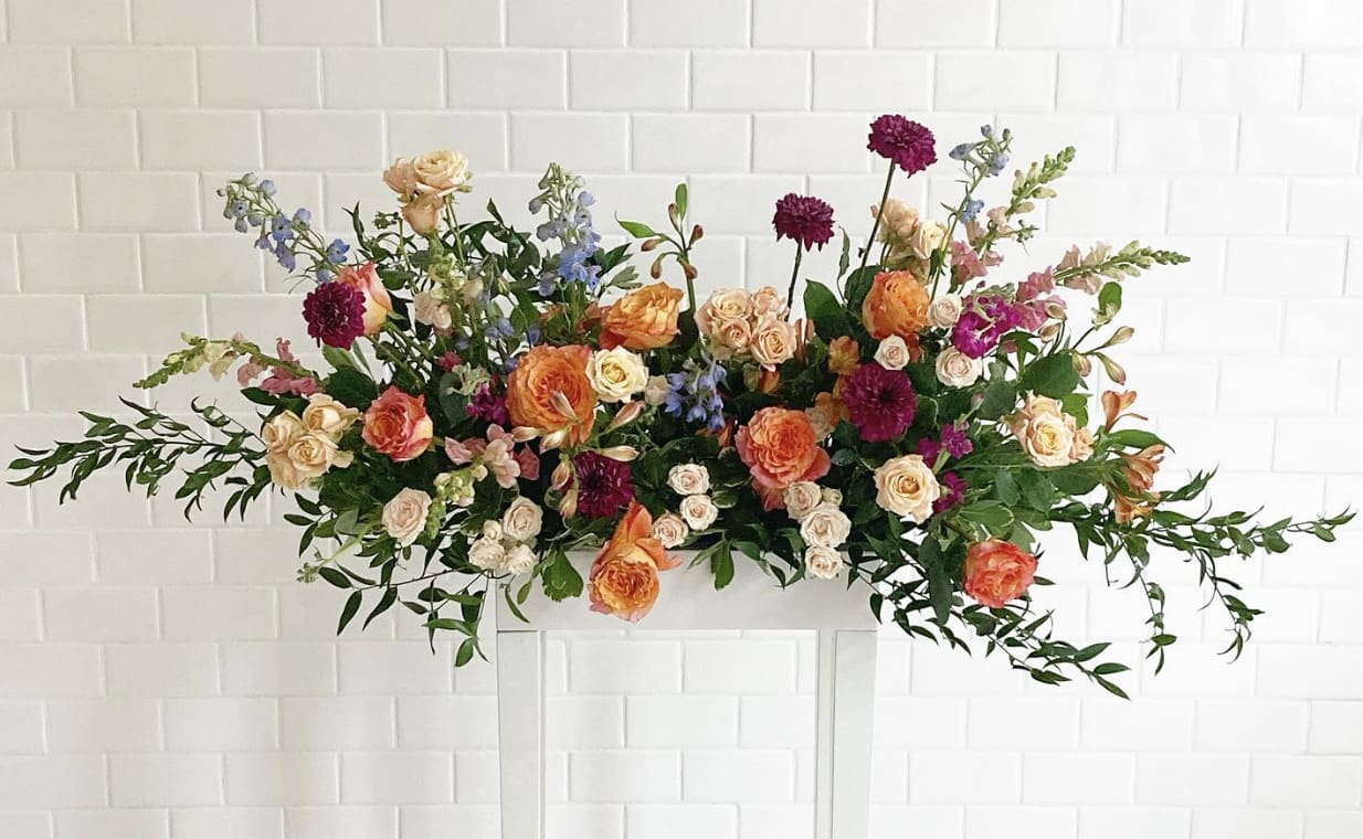 The Jade The Jade - Free spirit roses and dark purple mums create a captivating contrast, while cream spray roses offer a touch of elegance. Vibrant fuchsia stock and pink snapdragons bring lively color, softened by airy light blue delphinium and bright orange alstroemeria. Designed to be long and low, the Jade adds sophistication and cheer to any setting, perfect for making every occasion unforgettable.