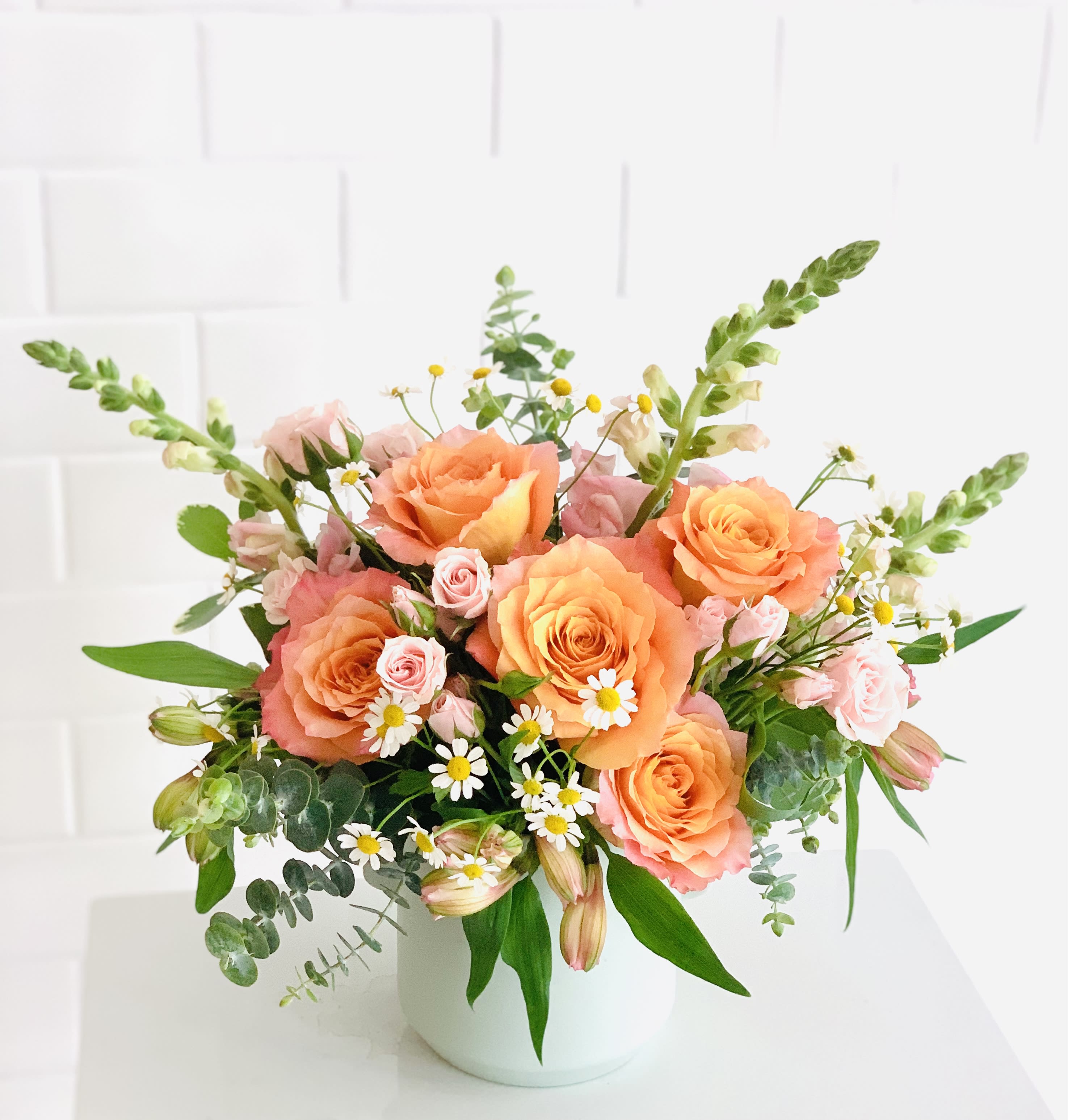 The Kierceton The Kierceton - Bring out your inner flower child with this whimsical arrangement. Free spirit roses are the star of the show with eye-catching bold beauty in contrast to a cool mint vase. Accompanied by snapdragons and spray roses, feverfew floats above them all to remind you to sway in the breeze and let your hair down every once and awhile.