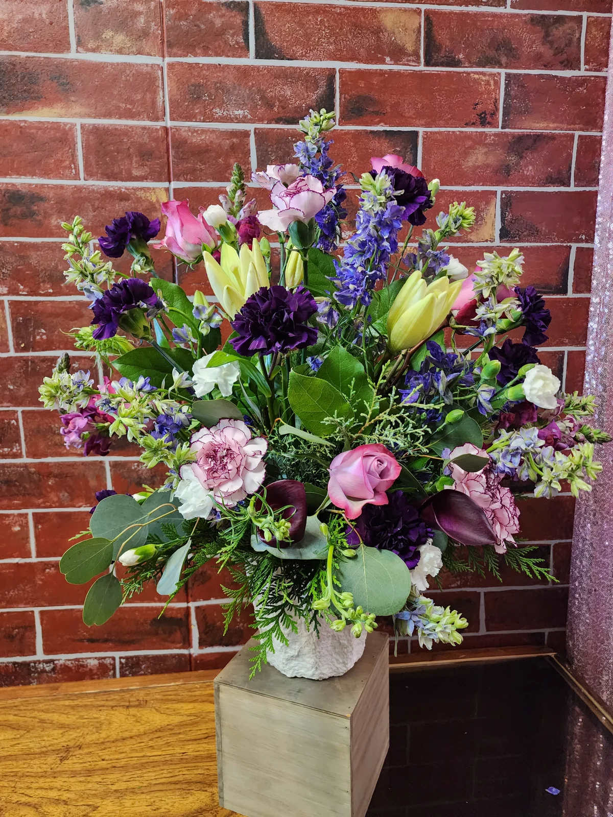 In Her Honor - A one-sided flower arrangement designed with fresh mix of flowers in shades of purple and white.  * Some stages of flower life may vary. (Example: Lilies may be more open than imaged.) Flower shortages and product availability means that some substitutions will apply. Value will always be filled. This bouquet might include eucalyptus amongst the greenery. It may be toxic to pets including but not limited to Cats and Dogs if digested.  Original Designer: Andrea Yager