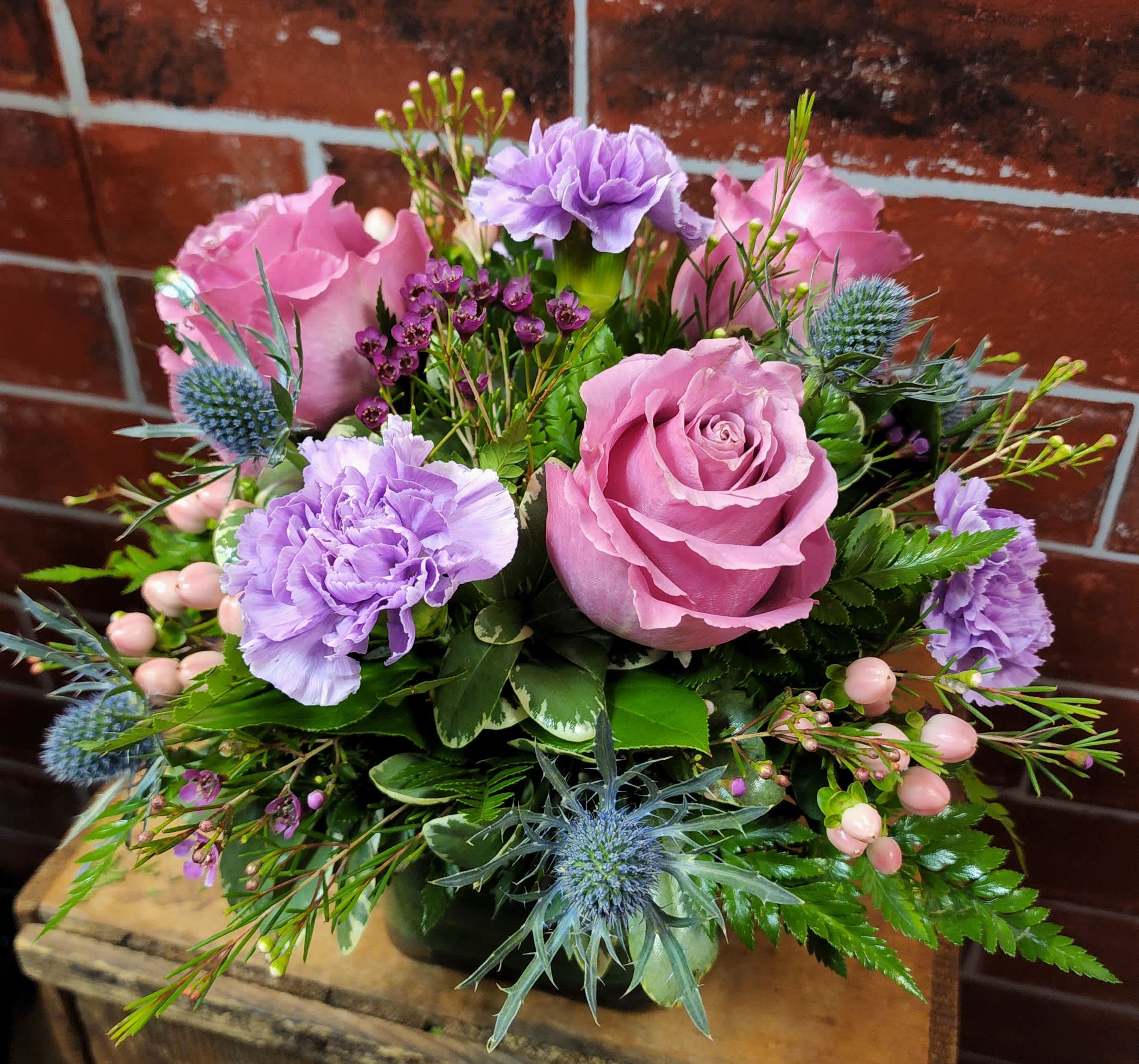 Lovely Square - This bouquet is in a square vase with a green leaf ribbon. Designed with purple carnations, pink hypericum, blue thistle, pink roses mixed with greens and filler flower.   * Some stages of flower life may vary. (Example: Lilies may be more open than imaged.) Flower shortages and product availability means that some substitutions will apply. Value will always be filled. This bouquet might include eucalyptus amongst the greenery. It may be toxic to pets including but not limited to Cats and Dogs if digested.
