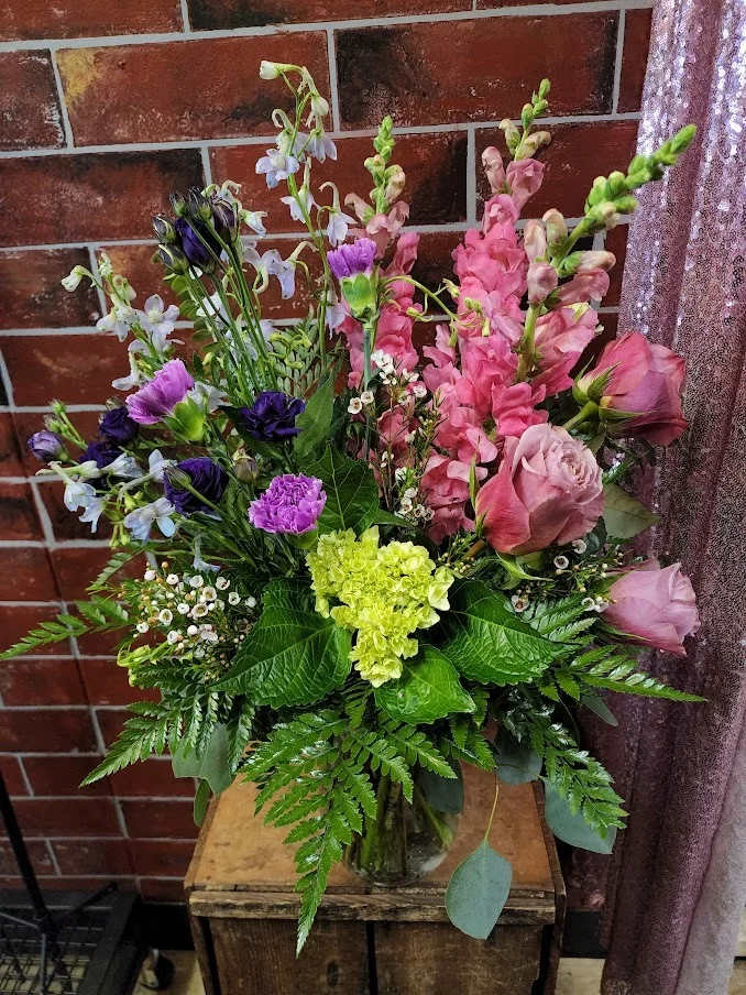 Pink, Purple &amp;  Blue - Designed with fresh flowers. This product is a one-sided design to include roses, snap dragons, carnations, blue delphinium, hydrangea and lisianthus.  * Some stages of flower life may vary. (Example: Lilies may be more open than imaged.)  Flower shortages and product availability means that some substitutions will apply. Value will always be filled.  This bouquet might include eucalyptus amongst the greenery. It may be toxic to pets including but not limited to Cats and Dogs if digested.