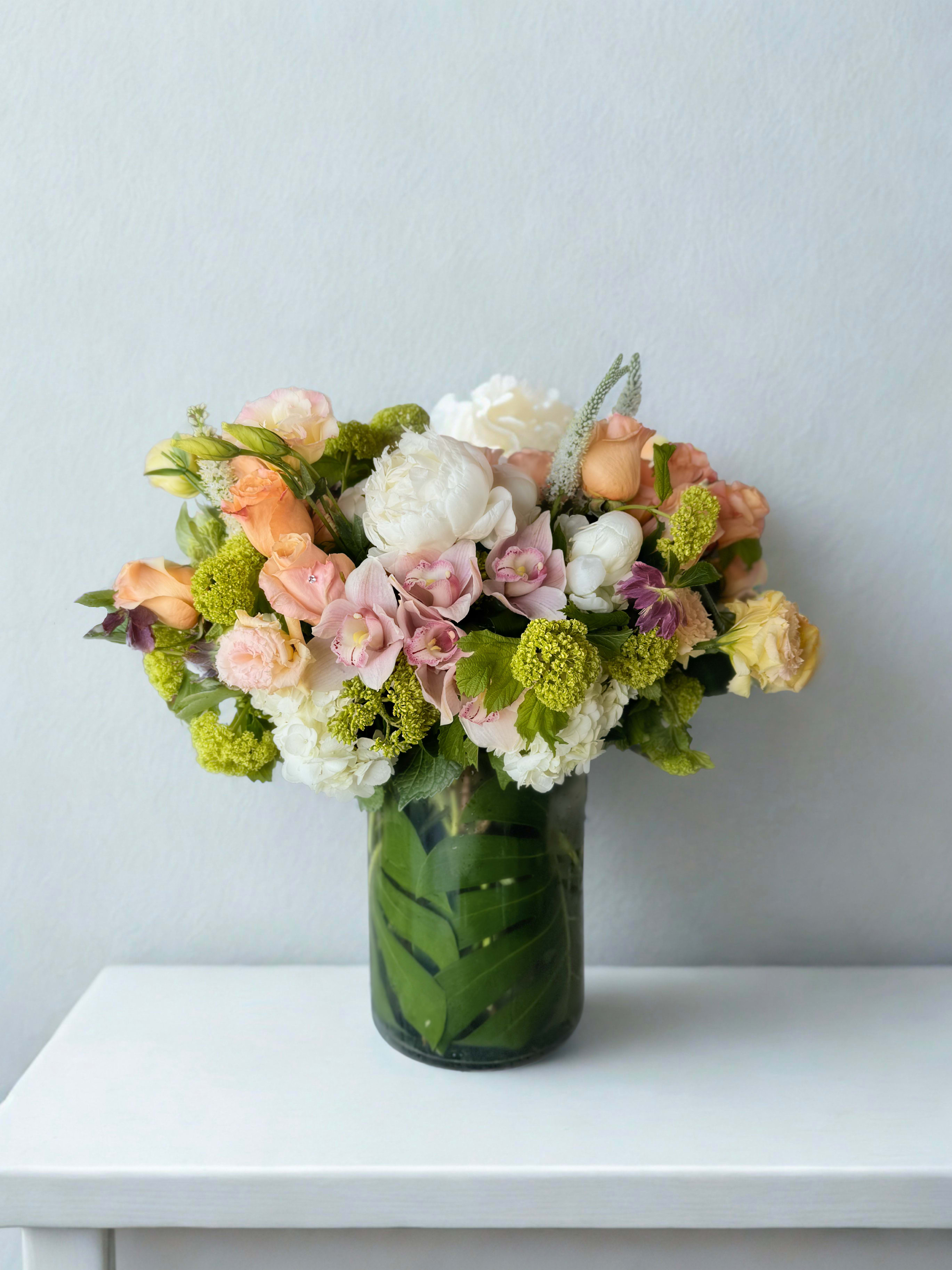 Peach Whisper - Grand Peonies, Rosies, Orchids - Ethereal and elegant, this arrangement is a delicate harmony of pastel beauty. Featuring creamy peonies, blush roses, soft pink cymbidium orchids, and textured green viburnum, among other blooms, it's all nestled in a chic glass cylinder lined with a modern leaf wrap. This bouquet exudes understated luxury and makes an ideal gift for weddings, anniversaries, baby showers, Mother's Day or simply to brighten someone’s space with refined charm.