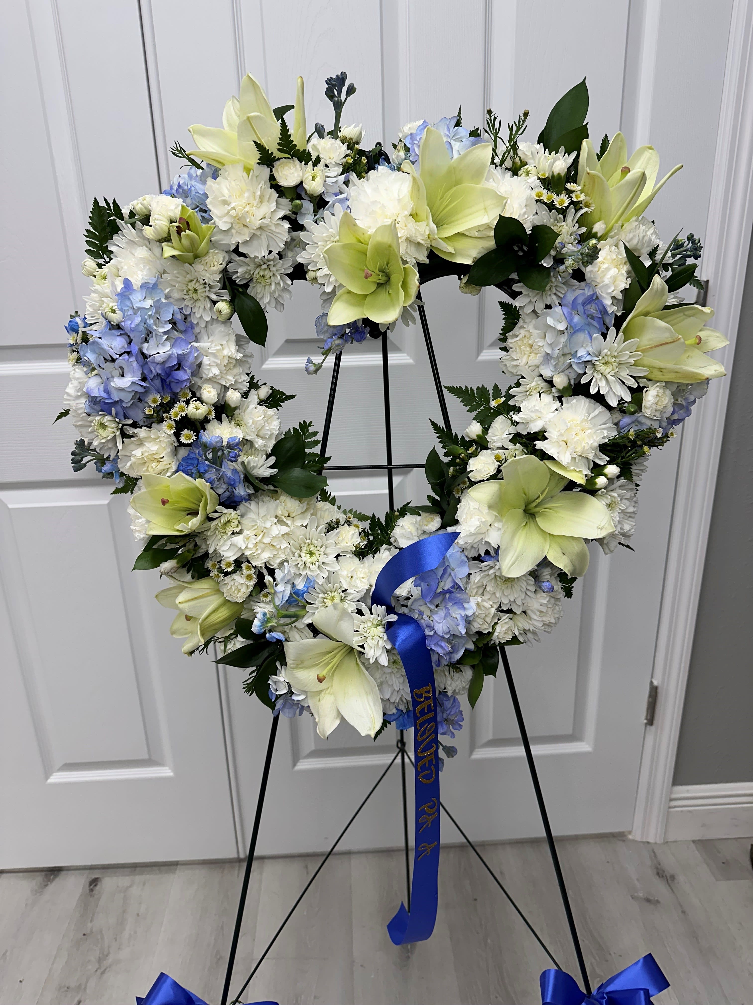 Solemn Embrace - Send a heartfelt embrace with our exquisite heart-shaped wreath, adorned with luscious white blooms of lilies and mums, delicately accented by the serene hues of blue hydrangea and stock. Each blossom whispers of love and remembrance, creating a poignant tribute to cherish. In this tender display, may the enduring spirit of your loved one be cradled in the gentle embrace of fond memories and everlasting affection.