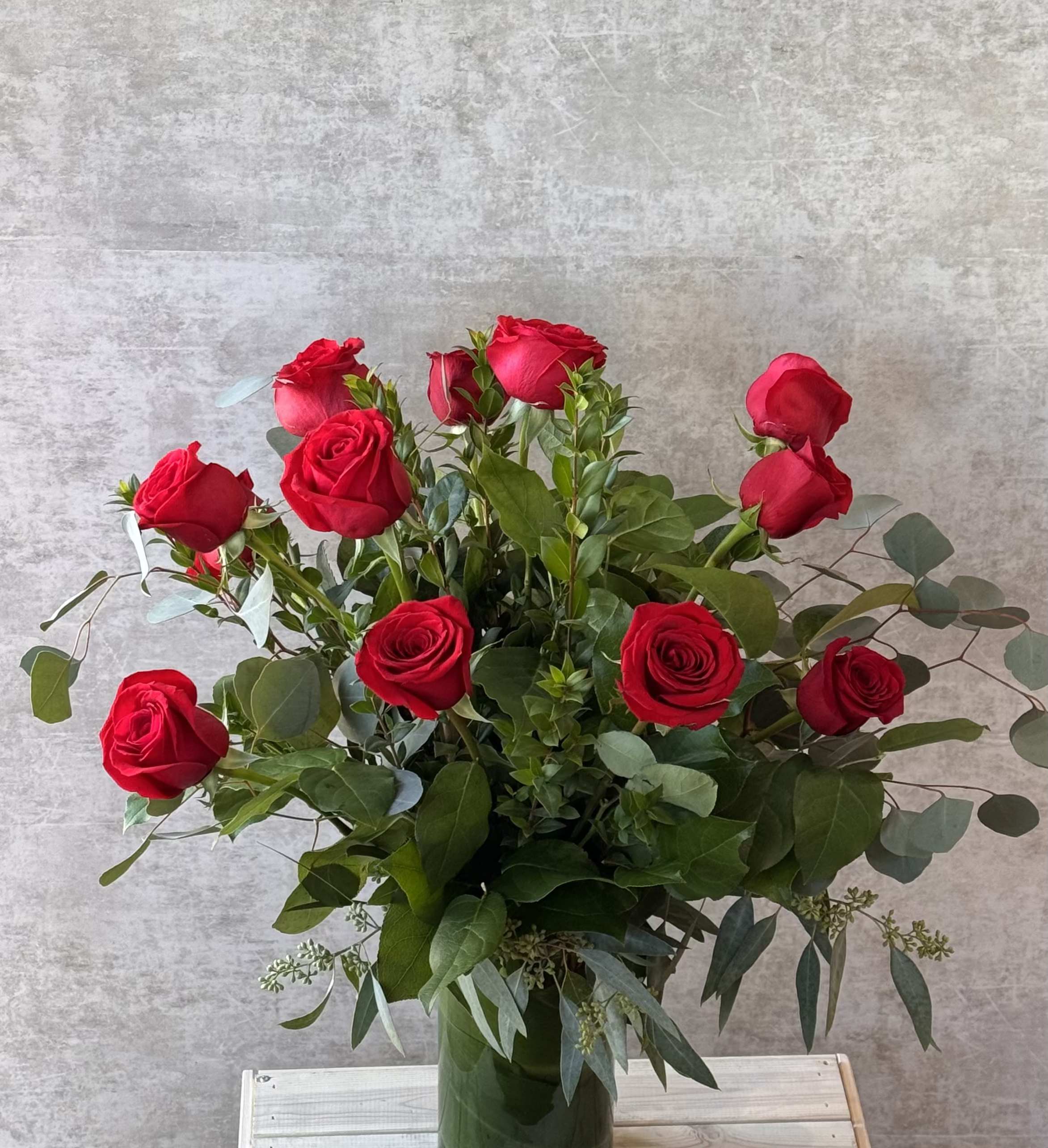 One Dozen Rose Arrangement One Dozen Rose Arrangement - One dozen rich red long stem roses elegantly arranged among lush eucalyptus and charming pink waxflower. This iconic arrangement, presented in a striking leaf lined glass vase, is the perfect symbol of love for Mother's Day, anniversaries, Valentine's Day, or any special moment.