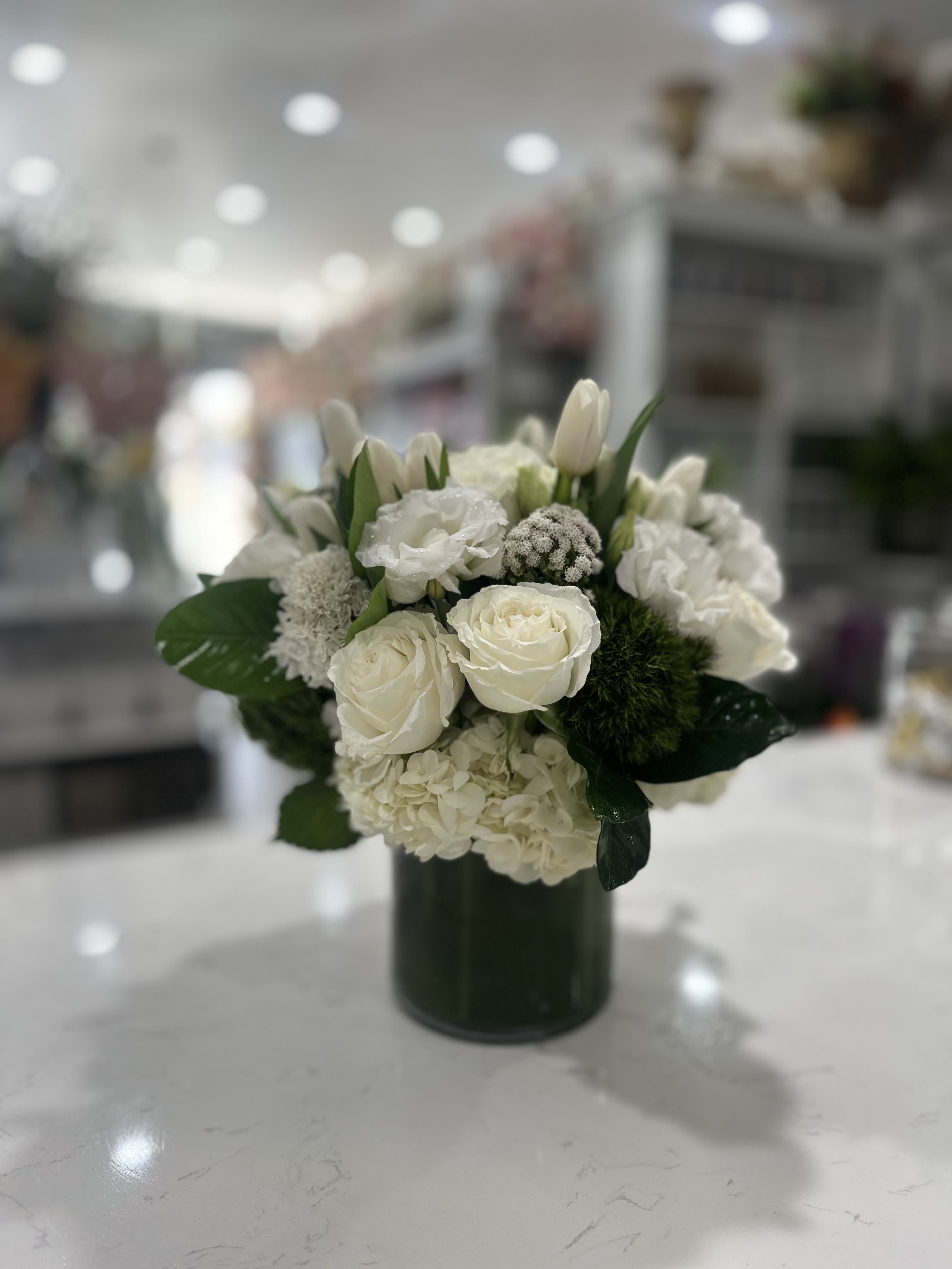 White Tranquility - An elegant, all-white floral arrangement featuring pristine roses, tulips, and hydrangeas, complemented by delicate green accents. Arranged in a classic glass vase, this piece exudes simplicity and grace, making it perfect for creating a serene atmosphere.