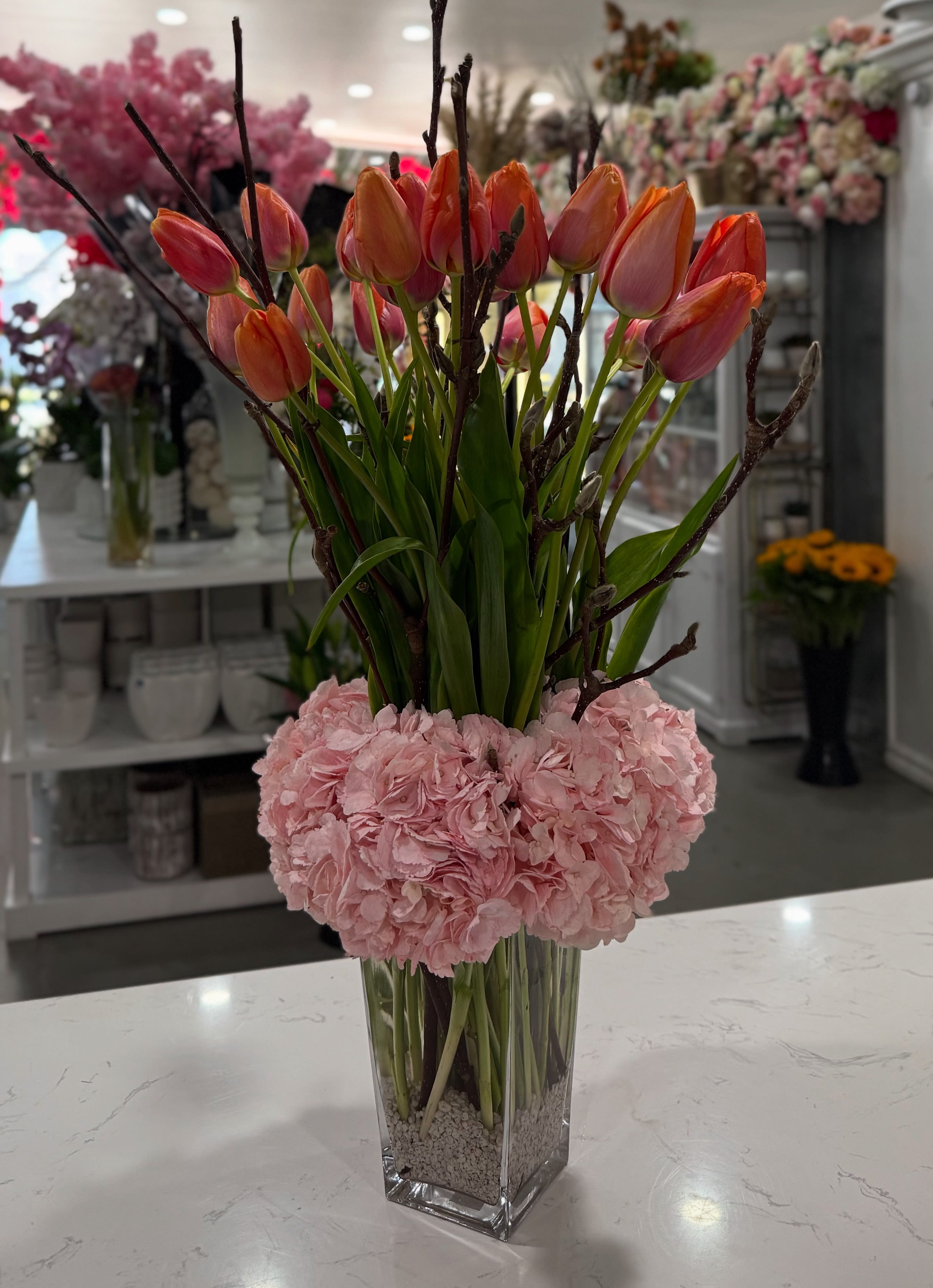Spring Ember - A striking arrangement of vibrant orange tulips rising above a cloud of soft pink hydrangeas, grounded in a clear vase with white pebbles. This bold yet elegant design captures the warmth of spring with a modern, architectural twist.
