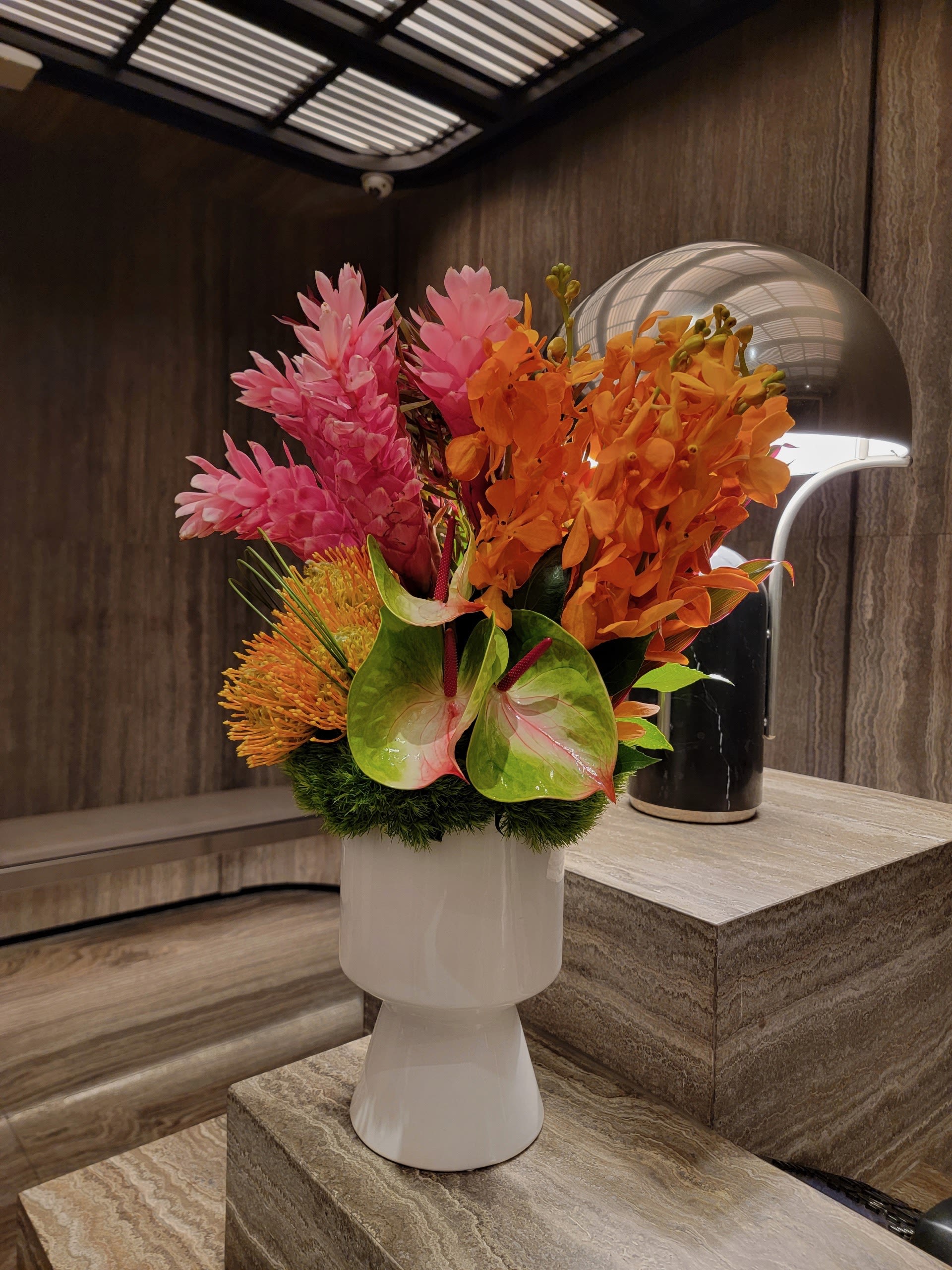 Tropical Muse - A bold and modern tropical arrangement featuring fiery orange orchids, pink ginger flowers, vibrant pincushions, and exotic anthuriums, all styled in a sleek white ceramic vase. Perfect for adding a pop of luxury and color to contemporary spaces.