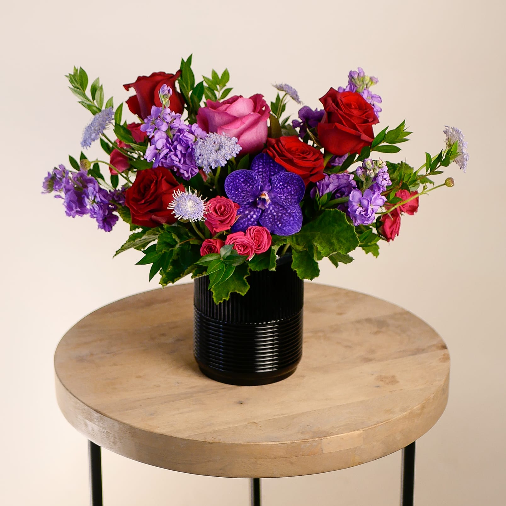 The Hideaway - This romantic arrangement features rich jewel tones of deep reds, fuchsia, and purples highlighting stunning roses and accents of fragrant stock and orchids in a moody black vessel. It exudes luxury and passion, making it the perfect choice for expressing love or celebrating a special moment.