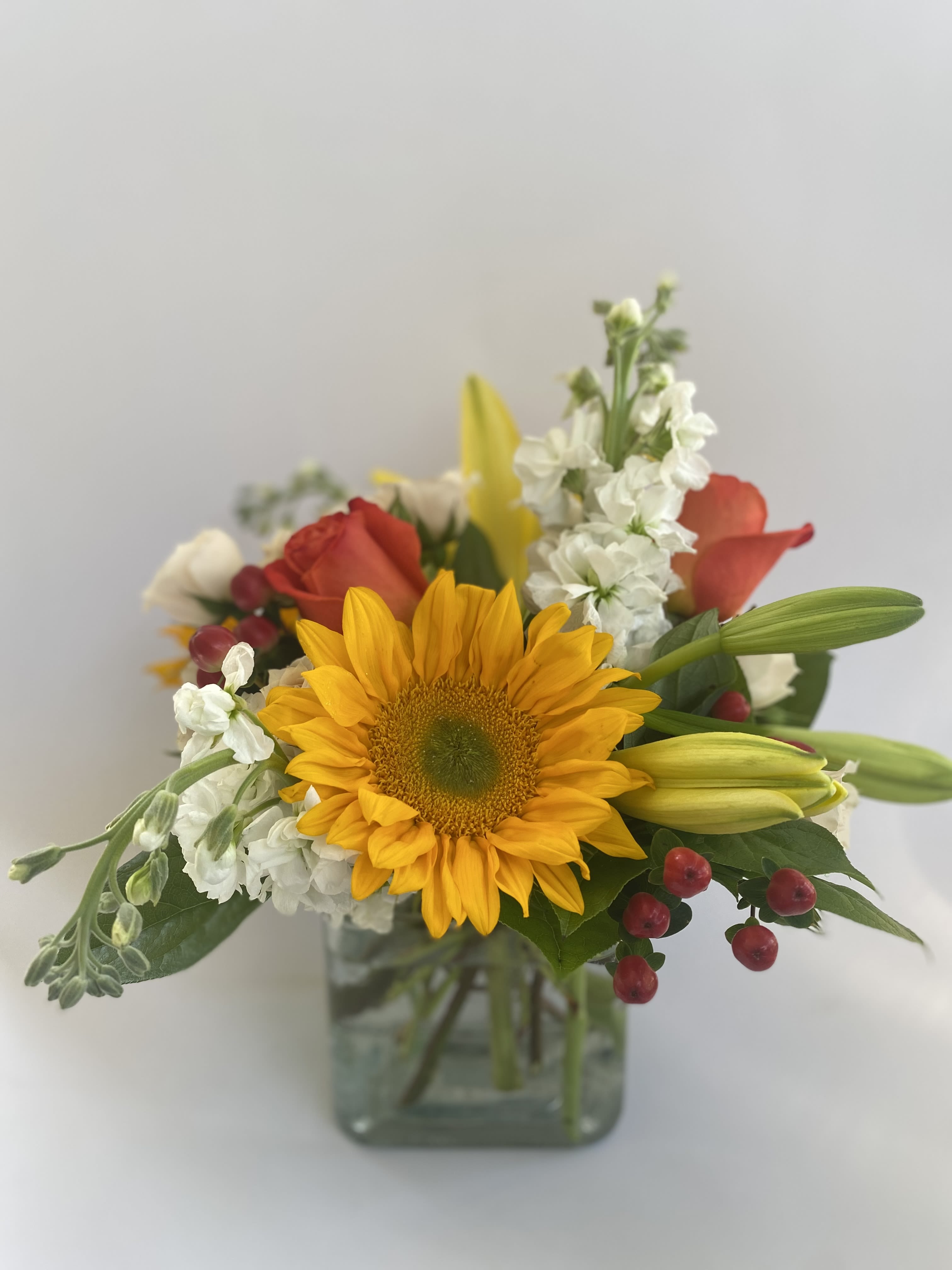 Seasonal Bliss - All the best of the season, this lovely mixture is a happy arrangement of today's favorite picks.  A modern square glass container is filled with seasonal florals such as spray roses, large roses, sunflowers, hypericum berries, stock, lilies and natural greenery. The standard size is pictured with options to upgrade to larger versions more flowers and a larger vessel. Seasonal flowers vary.