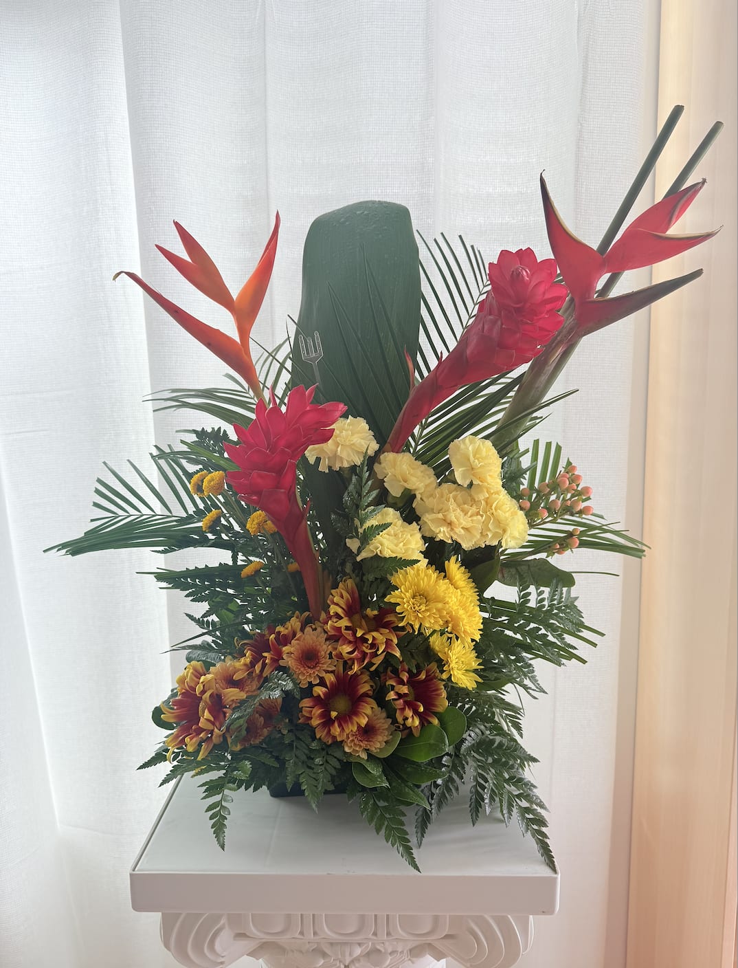 Tropical Grand Sunrise - Tropical Sunrise  Bring the warmth and vibrance of the tropics into any space with *Tropical Sunrise*. This bold arrangement features exotic heliconias, fiery ginger blooms, sunny yellow carnations, and daisy mums, all nestled among lush greenery. Designed to stand tall and proud, it's perfect for celebrating big milestones, expressing gratitude, or adding a striking touch to any room.  Lleva el color y la energía del trópico a cualquier espacio con *Tropical Sunrise*. Este arreglo llamativo combina heliconias exóticas, flores de jengibre vibrantes, claveles amarillos y margaritas entre follaje tropical. Ideal para celebrar logros, expresar agradecimiento o simplemente para impactar con un toque audaz y tropical. 