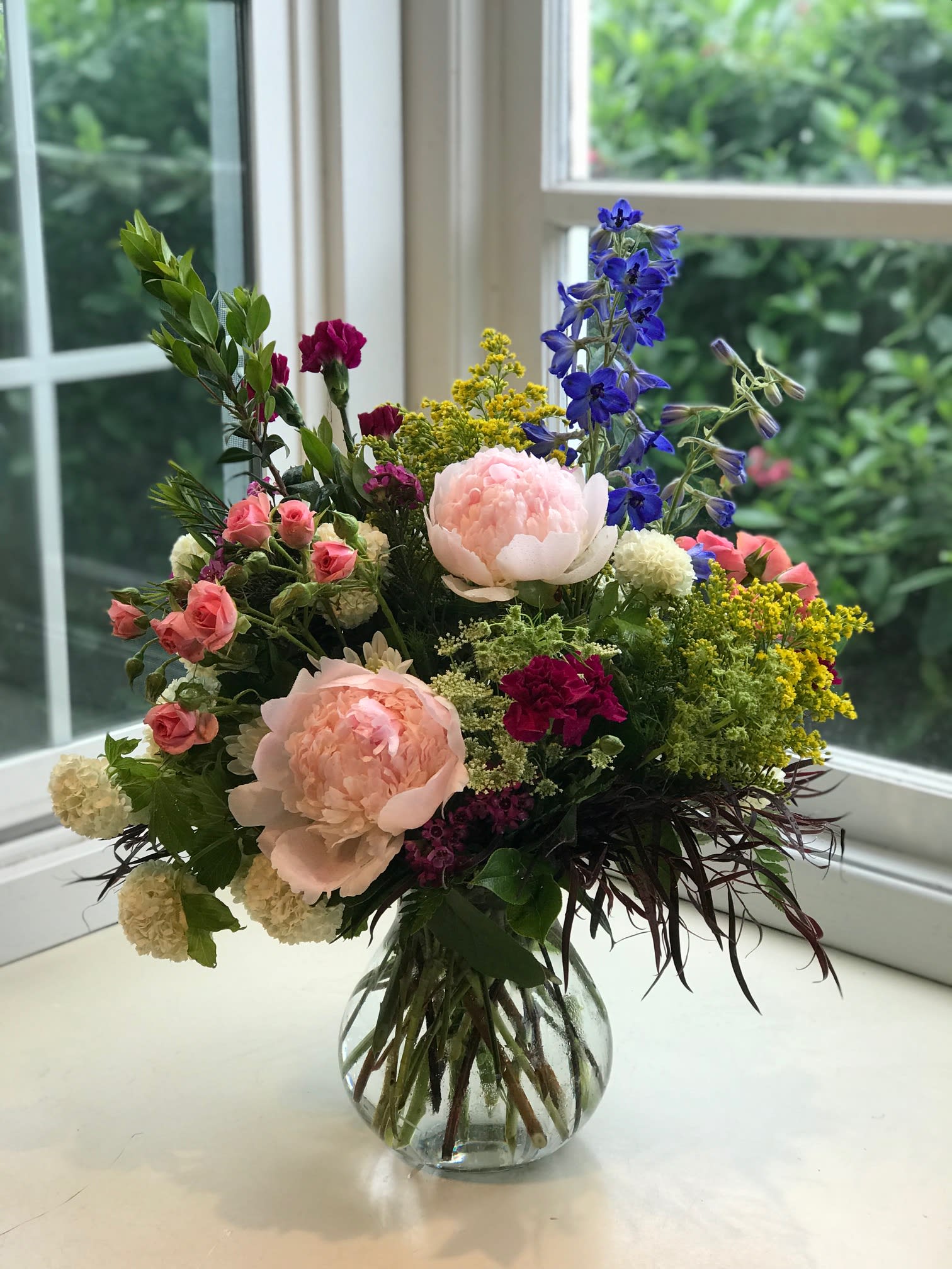 Ode to Spring - This bright and colorful design features lovely peonies (when in season,) a pop of  blue delphinium, hot pink spray roses, and a touch of yellow. As always, designer may substitute based on what we have in stock.  Call shop to get specifics. *Peonies are not currently available.