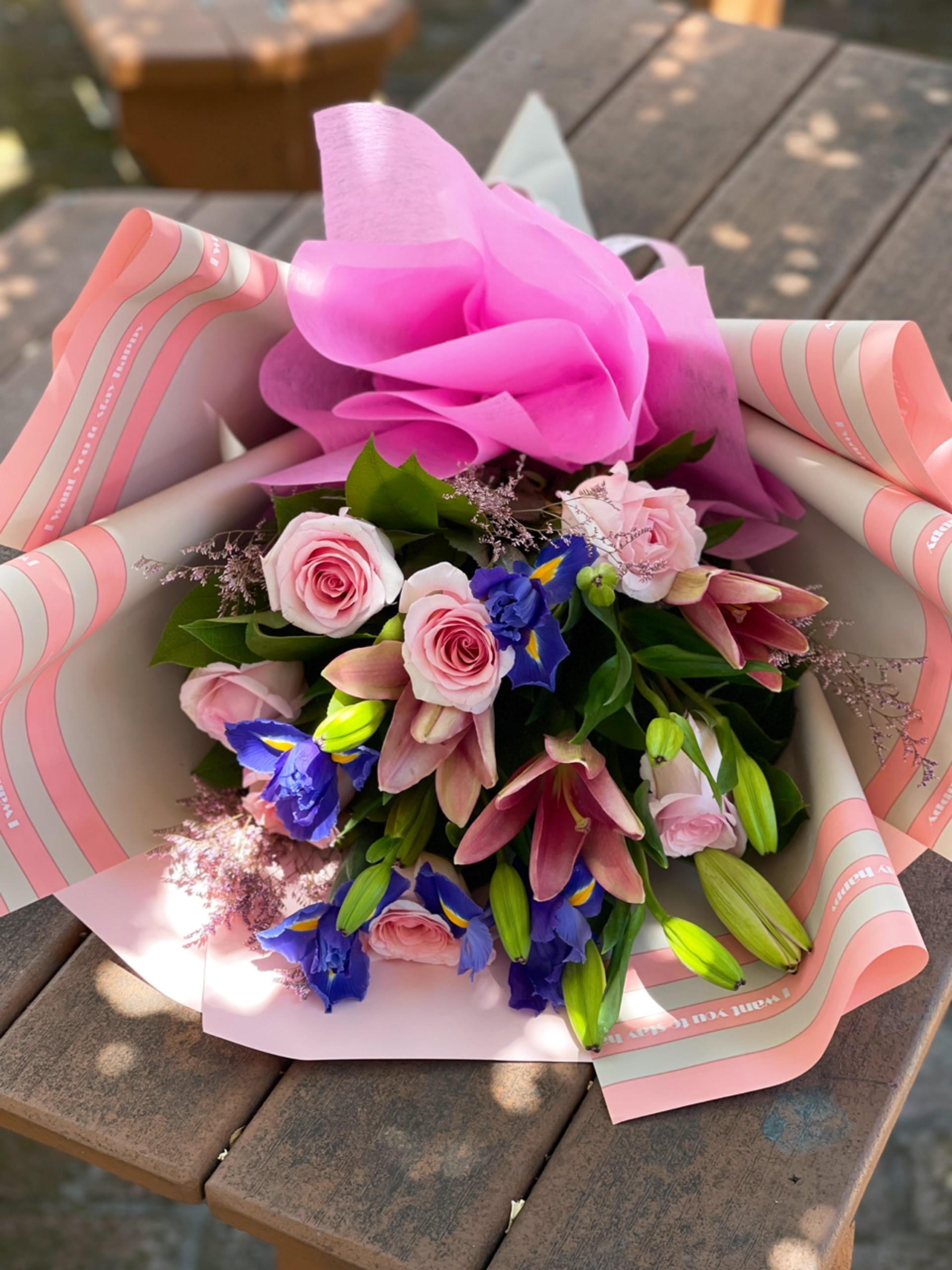 Pink Roses & Iris Wrap Bouquet Pink Roses & Iris Wrap Bouquet - - 8 Pink Roses - 4 Blue Iris - 3 Stems Pink Lilies - Fillers - Greenery - Paper and Bow