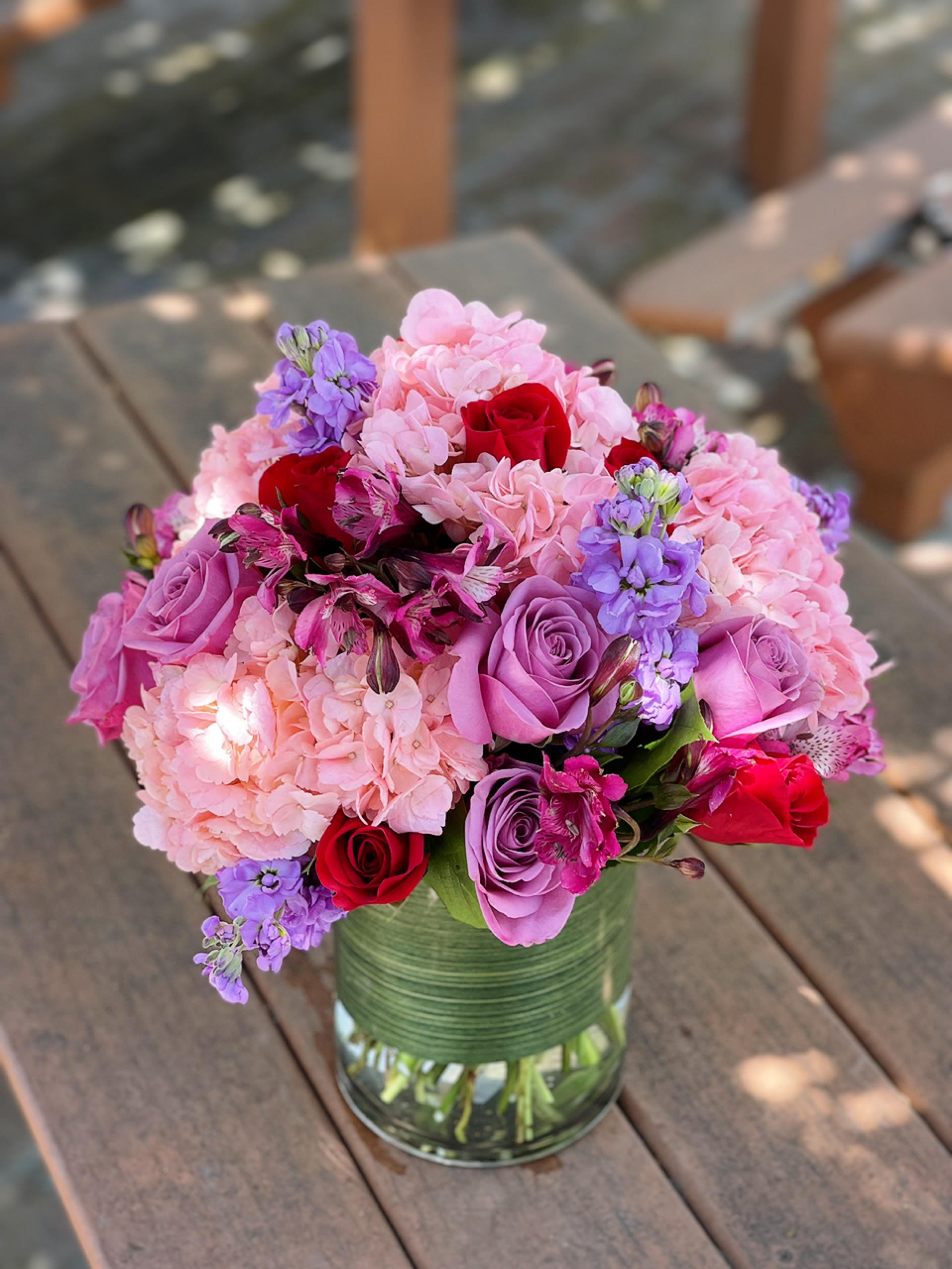 New Day - - 4 Pink Hydrangeas - 5 Lavender Roses - 5 Red Roses - 5 Purple Stock - 4 Purple Alstroemeria - Greenery - 6x8 Inches Cylinder Vase
