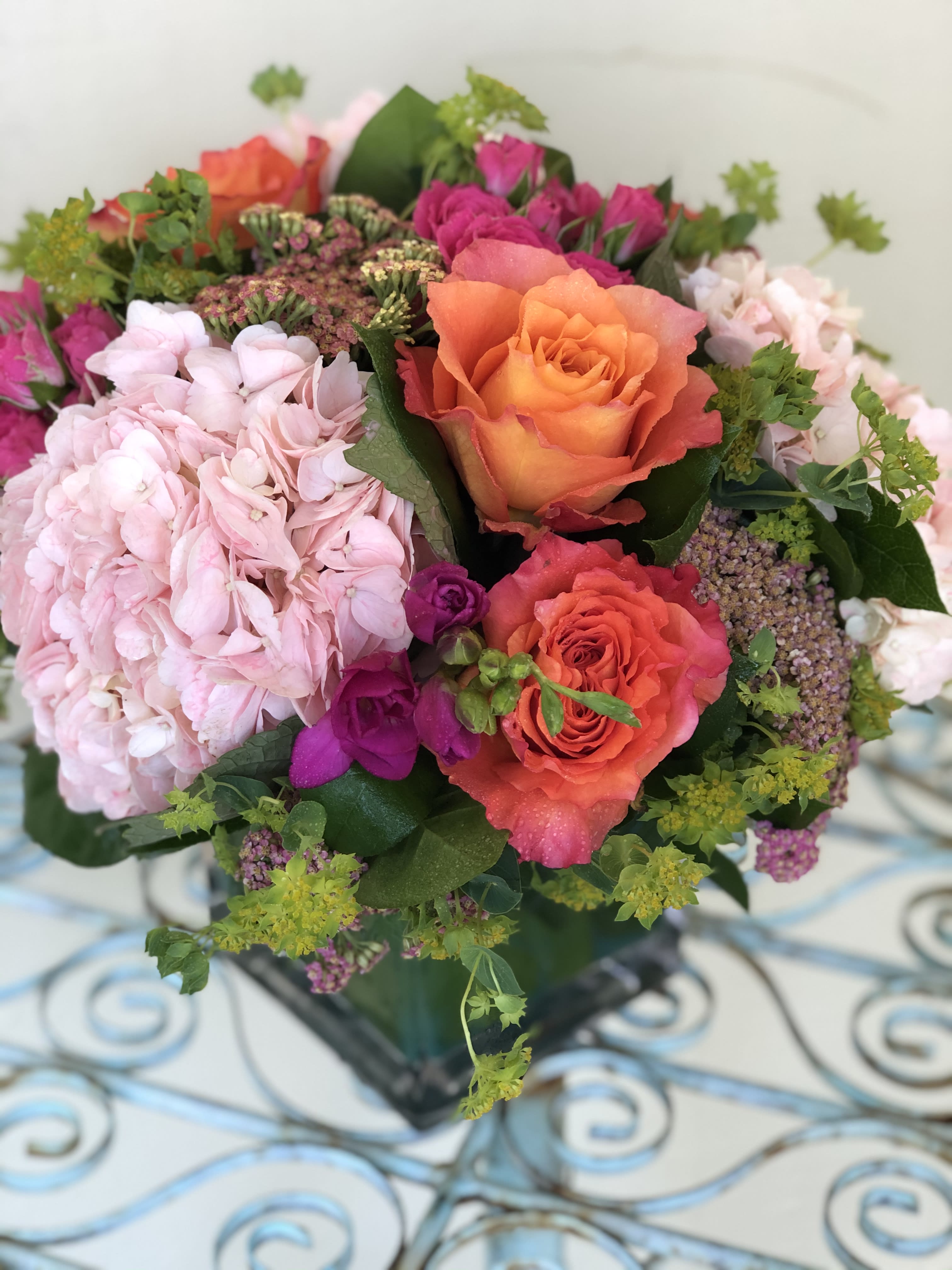 Sunrise - Cheerful combination Orange and pink seasonal flowers including Hydrangeas &amp; Roses in a 5&quot; cube lined with Ti leaves. The deluxe will be in 6x6 glass cube. Flowers are subject to change due to seasonal availability, however we will create a similar arrangement.