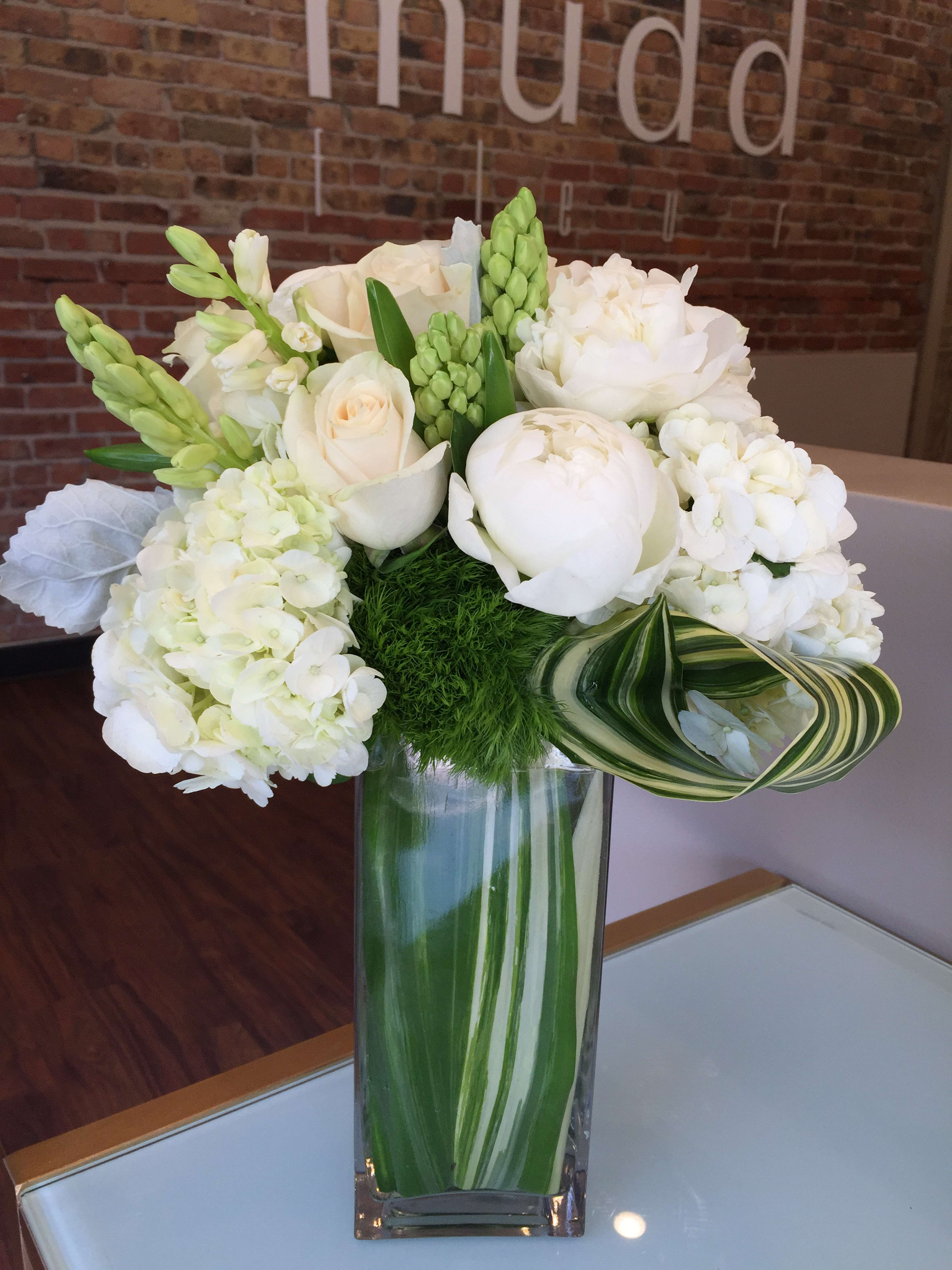 Designers Pick of Premium Seasonal White and Green Blooms will fit in any space! Designers Pick of Premium Seasonal White and Green Blooms will fit in any space! - A designers choice, classic bouquet of premium white and green flowers in a 12" tall rectangular glass vase lined with a leaf wrap and accented with rolled foliage. Flower varieties will vary depending on market availability and arrangement will not be exactly as pictured but still in the same style.