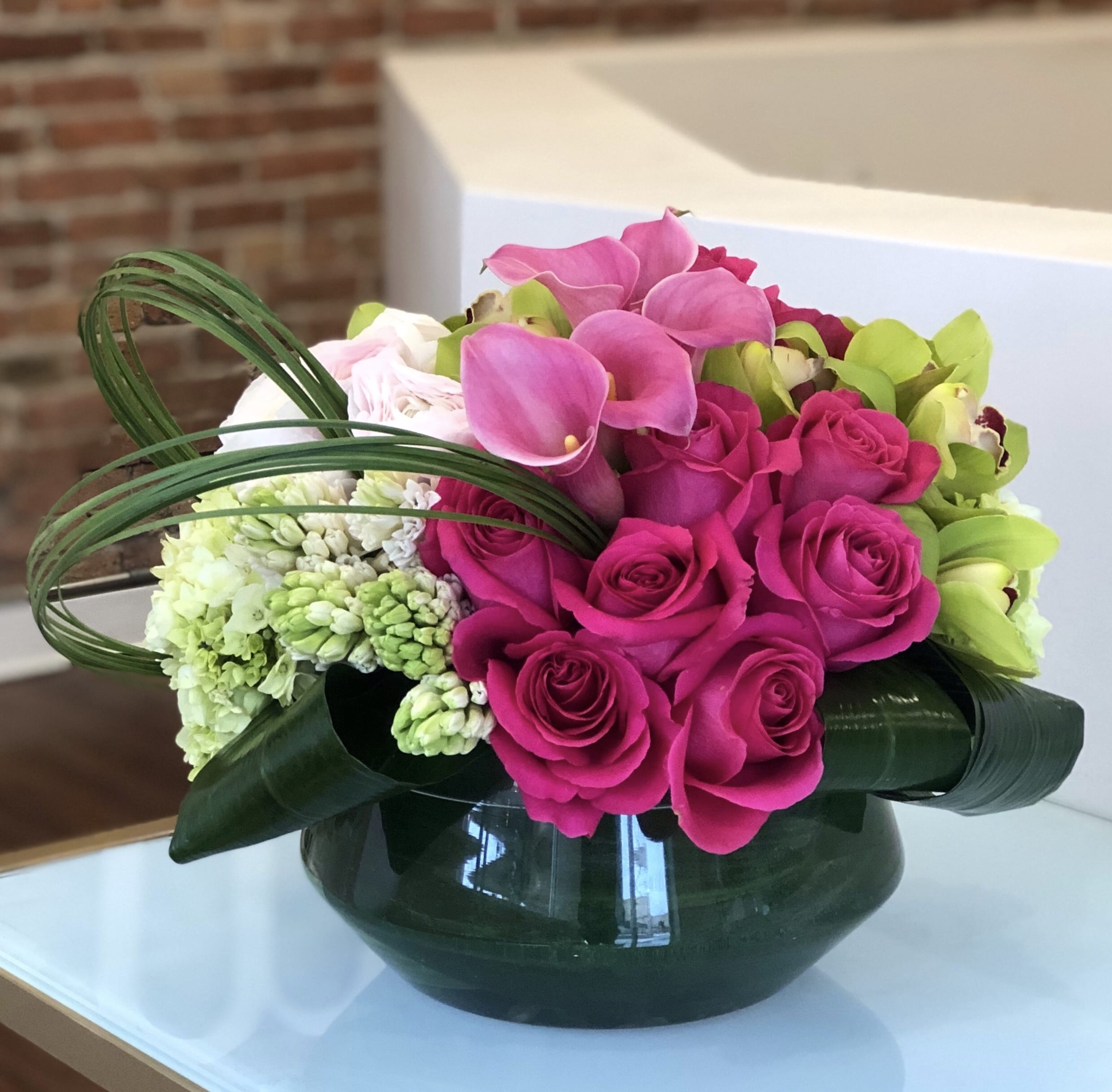Designers Choice Spring Garden Designers Choice Spring Garden - A designers choice arrangement of bright and light pink flowers along with shades of bright green for a bold spring pop. Flower varieties will vary depending on market availability and will not be as pictured but designed in the same style. Pictured flowers include bright pink calla lilies and roses with blush hyacinth and ranunculus and lime green hydrangea and cymbidium orchids designed in a low clear glass bowl with a leaf wrap and bear grass loops.