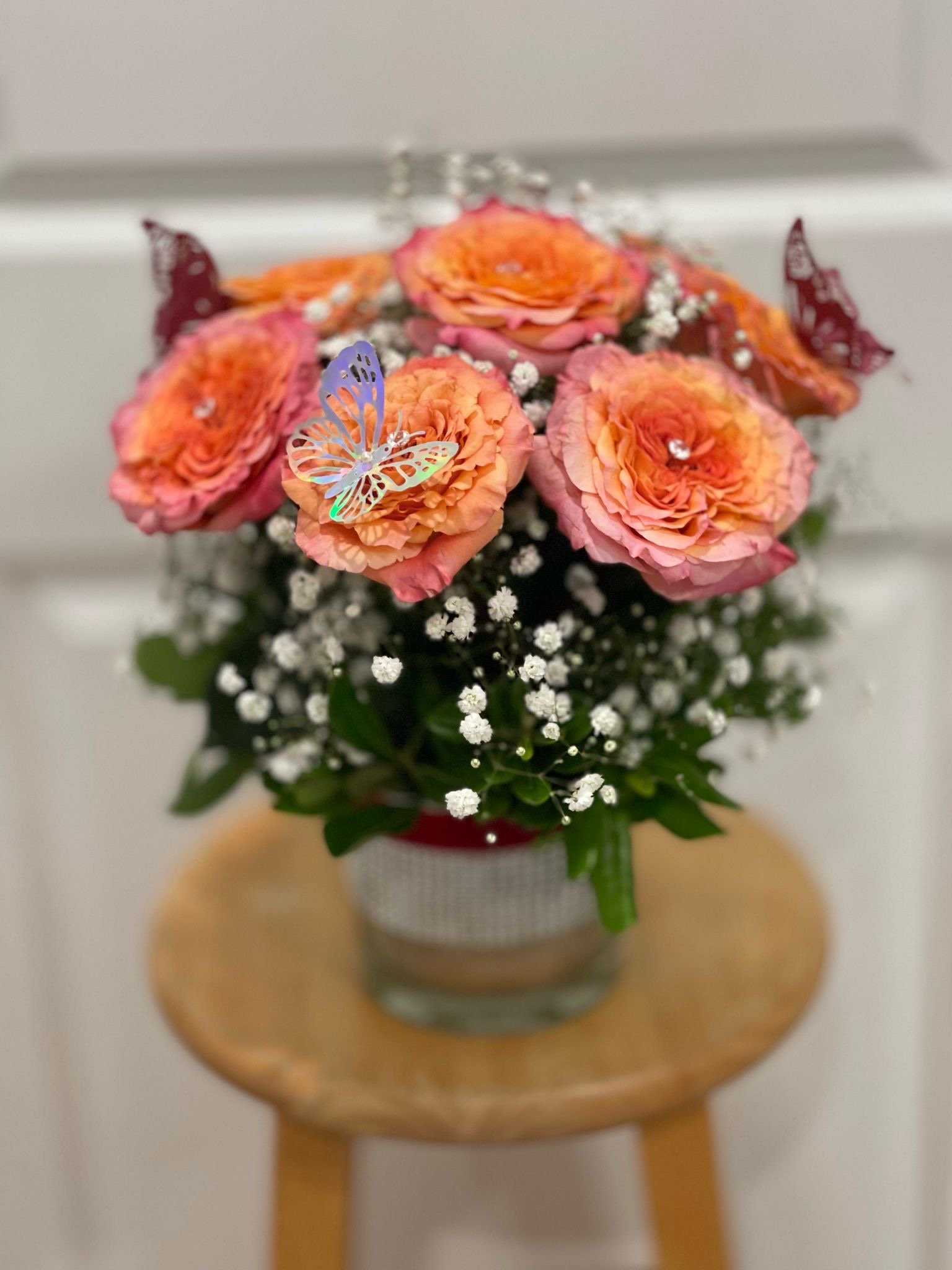 Sunset Whispers Butterfly Rose Vase Sunset Whispers Butterfly Rose Vase - Sunset Whispers Butterfly Rose Vase Let your sentiments bloom with our “Sunset Whispers” Butterfly Rose Vase, a charming arrangement that captures the warmth of golden hour. Featuring peachy-orange garden roses with hints of blush, nestled in a bed of dainty baby’s breath and fresh greenery, this bouquet brings a gentle touch of magic to any space. Topped with iridescent and burgundy butterflies and subtle jewel accents, it’s a playful yet elegant design—perfect for birthdays, thank-you gifts, or simply brightening someone's day. Details: - 6 premium peach ombré roses - Baby’s breath and greenery for soft texture - Glass vase with rhinestone ribbon detail - Butterfly embellishments for a whimsical touch - Ideal for heartfelt everyday gifting or cheerful decor A bouquet that feels like a hug, kissed by butterflies and sunshine.