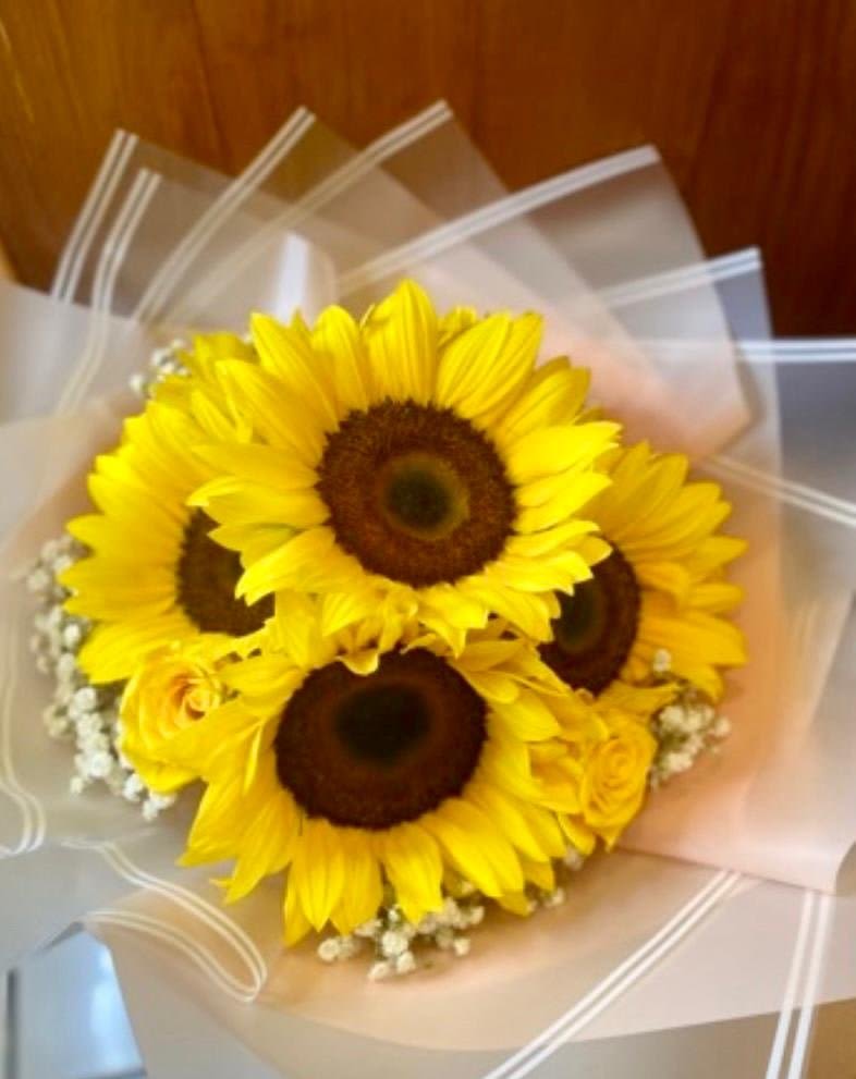 Sunshine Blossom Bouquet* Sunshine Blossom Bouquet* - Sunshine Blossom Bouquet Brighten someone's day with our Sunshine Blossom Bouquet, a cheerful arrangement that exudes warmth and joy. This beautiful bouquet features four vibrant sunflowers, their golden petals symbolizing happiness and positivity, paired perfectly with the soft elegance of yellow roses. The delicate white baby’s breath adds a touch of grace and freshness, while the unique Japanese paper wrapping brings an artistic and refined finishing touch to this radiant arrangement. Whether it's a birthday, a celebration, or just to spread a little sunshine, this bouquet is sure to brighten any occasion with its joyful colors and timeless charm. Bright. Elegant. Uplifting. A perfect gift to share a little light and love.