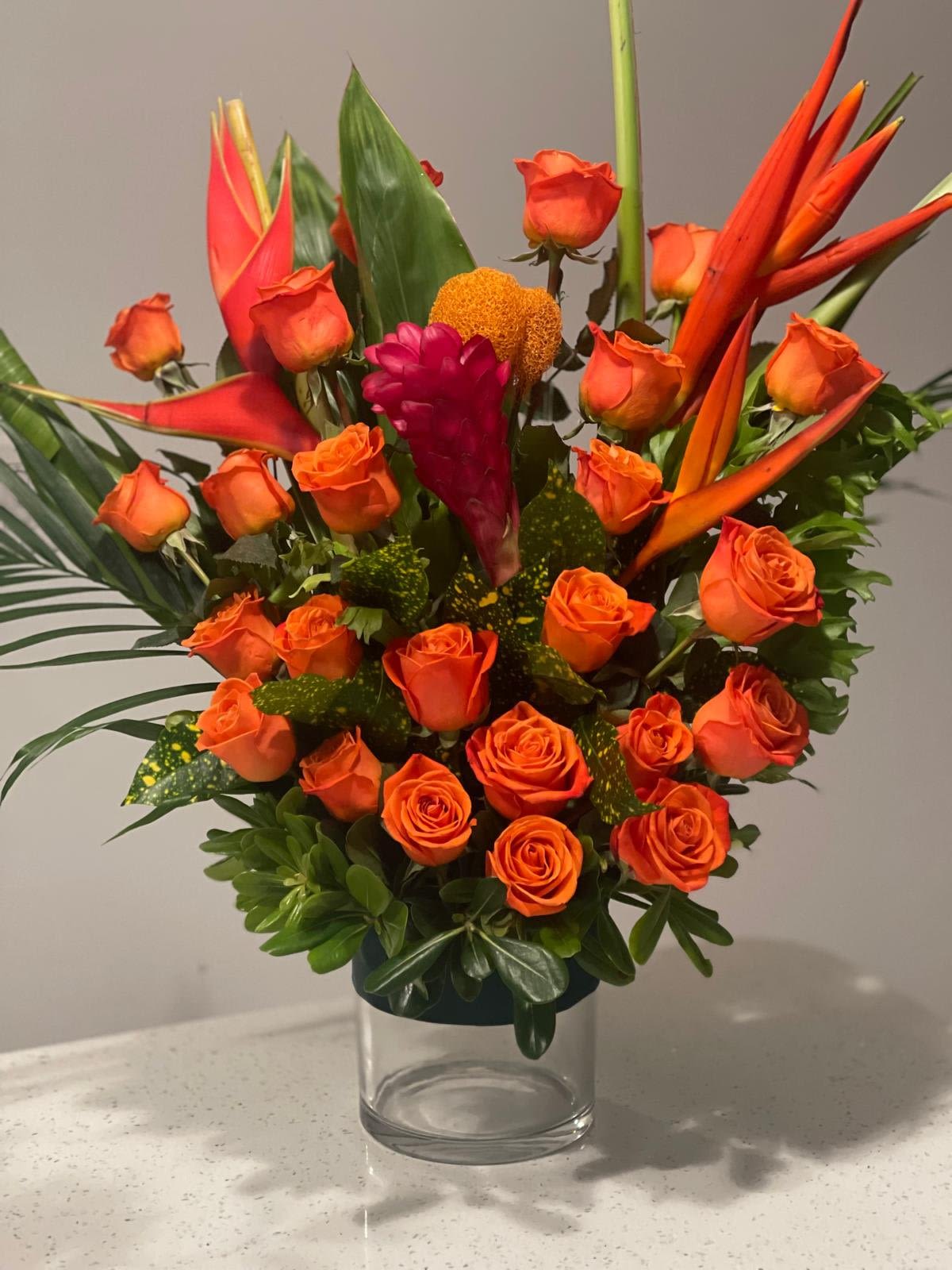 XL Tropical Romance Arrangement XL Tropical Romance Arrangement - Indulge in the bold beauty of our XL Tropical Romance Arrangement. This lavish design brings the vibrant hues of nature into any space, featuring a striking blend of orange roses, lush greenery, exotic Birds of Paradise, and elegant ginger flowers. With an array of carefully chosen tropical greenery, this arrangement is a true celebration of the rich colors and textures of the tropics. Presented in a sleek, rounded glass container, the XL size makes a dramatic statement, perfect for creating a luxurious focal point in any room. The dynamic mix of tropical blooms and foliage evokes a sense of romance and adventure, making it an unforgettable gift for any occasion. Bold. Exotic. Unforgettable. Bring the beauty of a tropical paradise into your home with this stunning XL arrangement.