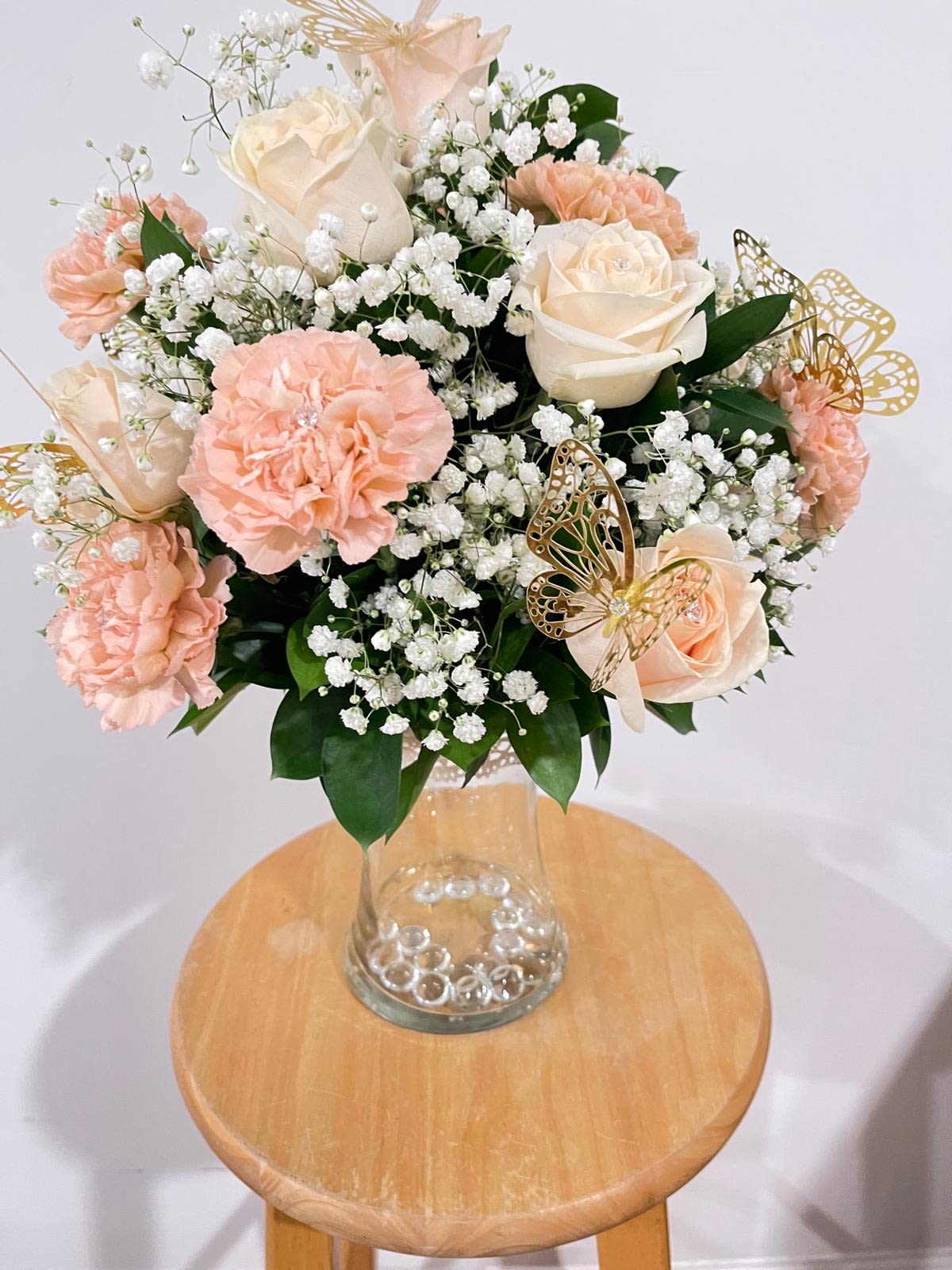 &quot;Blushing Garden” Rose &amp; Carnation Bouquet - Float away into a world of delicate beauty with our Blushing Garden Rose &amp; Carnation Bouquet**. A graceful pairing of creamy ivory roses, peachy-pink carnations, and clouds of baby's breath, this arrangement is finished with lush greenery and sparkling golden butterfly accents. Each bloom is delicately kissed with a crystal center, adding a subtle shimmer to this enchanting design.  Perfect for birthdays, anniversaries, baby showers, or simply brightening someone’s day, this bouquet feels like a tender embrace in floral form.  Details - Soft neutral and peachy tones for a sophisticated look   - Premium roses, carnations, and baby's breath   - Elegant gold butterfly accents and rhinestone embellishments   - Stylish clear glass vase with bubble accents   - Ideal for: Celebrations, sympathy, new beginnings, or romantic gestures   A bouquet where elegance meets pure magic, one petal at a time. 