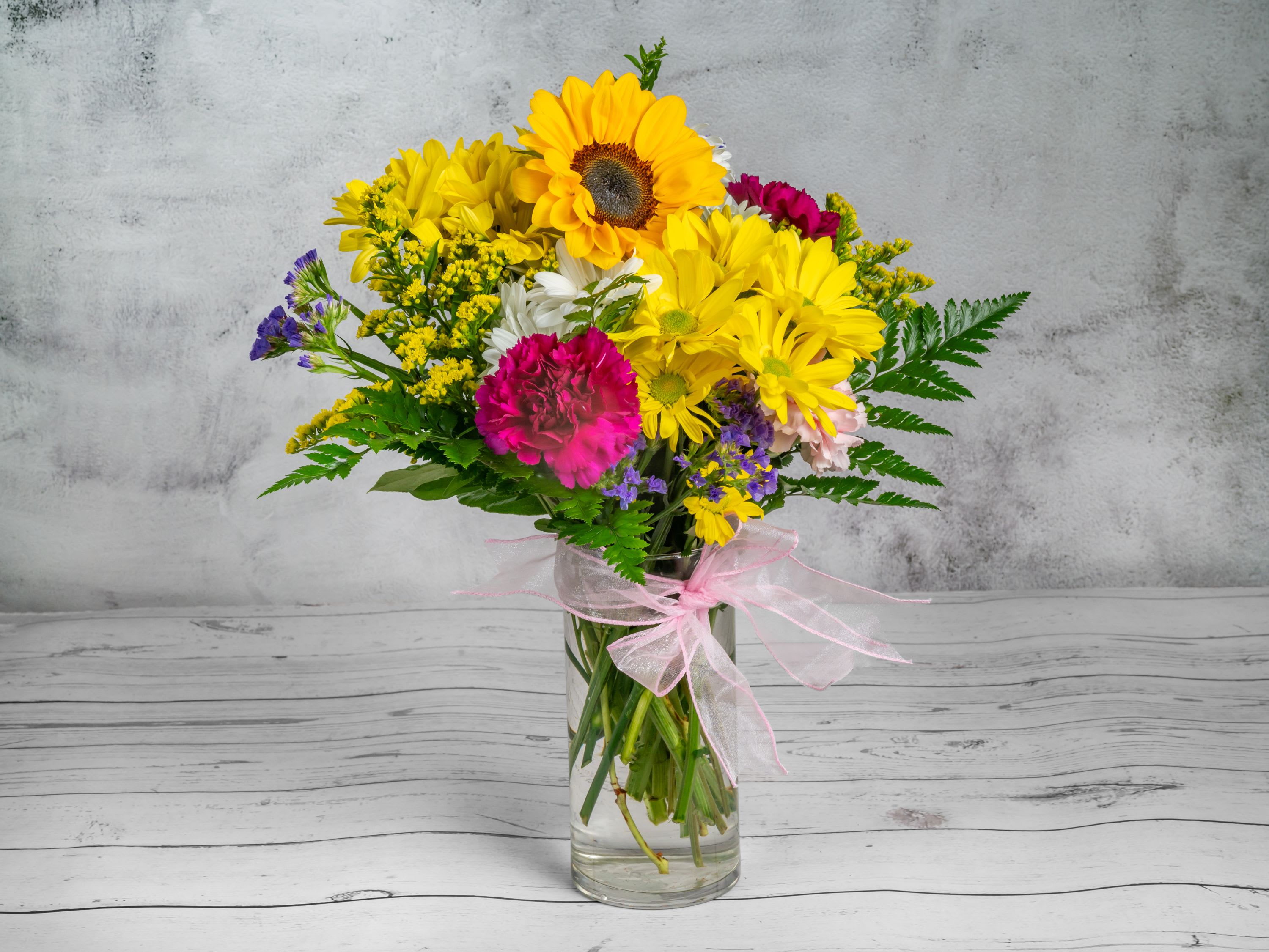 Happy Blooms Happy Blooms - Brighten any room with this cheerful and vibrant flower arrangement! This bouquet features a stunning mix of sunflowers, yellow daisies, pink carnations, and delicate purple accents, complemented by lush green foliage. Arranged in a clear glass vase tied with a soft pink ribbon, this display exudes warmth and joy, making it perfect for birthdays, celebrations, or simply as a thoughtful gesture to brighten someone's day. The rustic yet elegant design fits seamlessly into any décor, spreading happiness wherever it's placed.