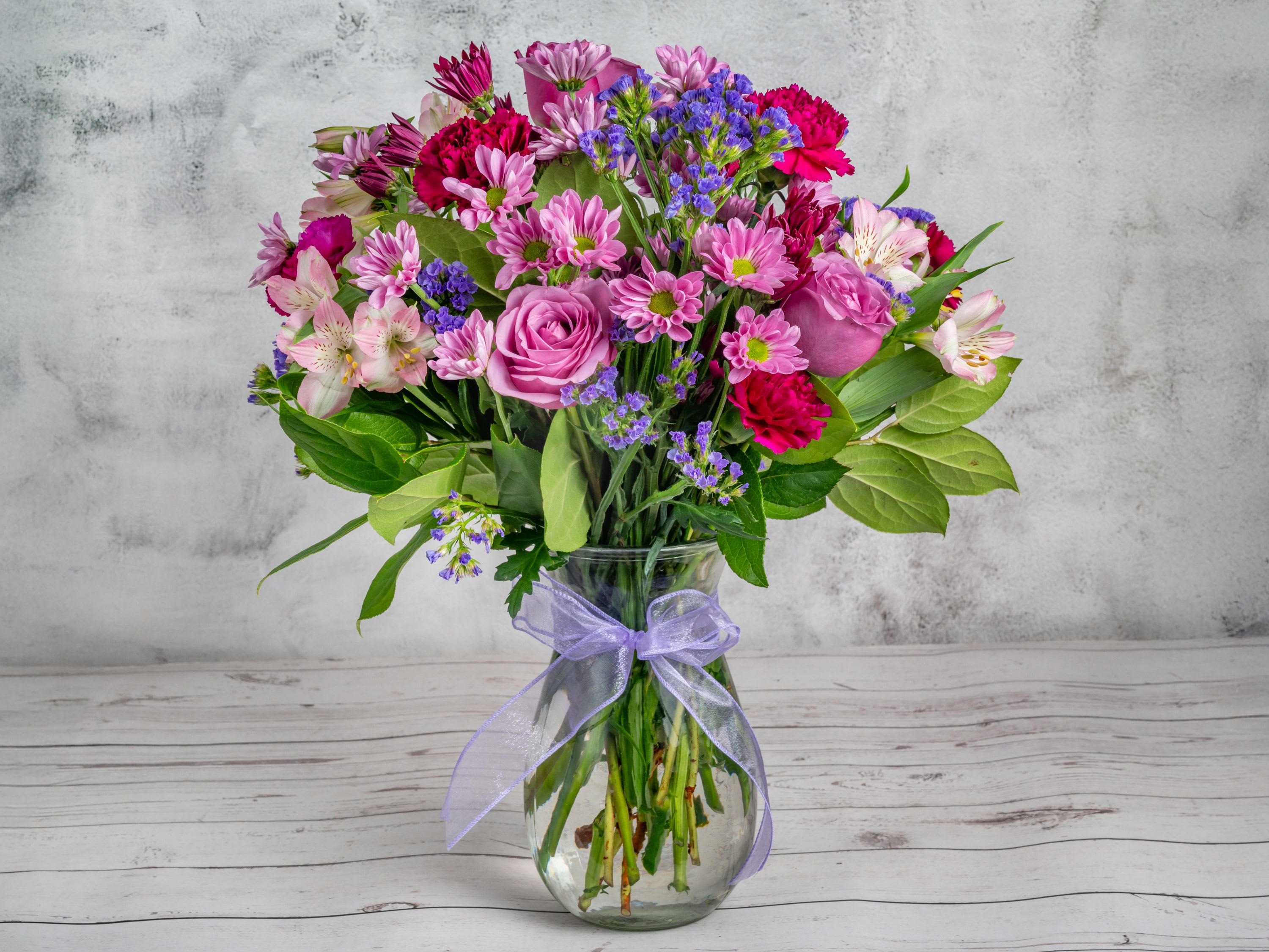 Lavender Elegance Lavender Elegance - This enchanting bouquet features a delicate blend of soft lavender roses, lavendar daisies, and striking magenta carnations and alstroemerias. Accents of lush greenery and sprigs of deep purple statice add depth and texture to this harmonious display. Nestled in a classic glass vase adorned with a sheer lavender ribbon, this arrangement is the perfect choice for expressing admiration, celebrating milestones, or simply bringing joy to someone’s day. Each bloom is carefully selected for its vibrant color and freshness, ensuring that this bouquet stays radiant and lovely for as long as possible.