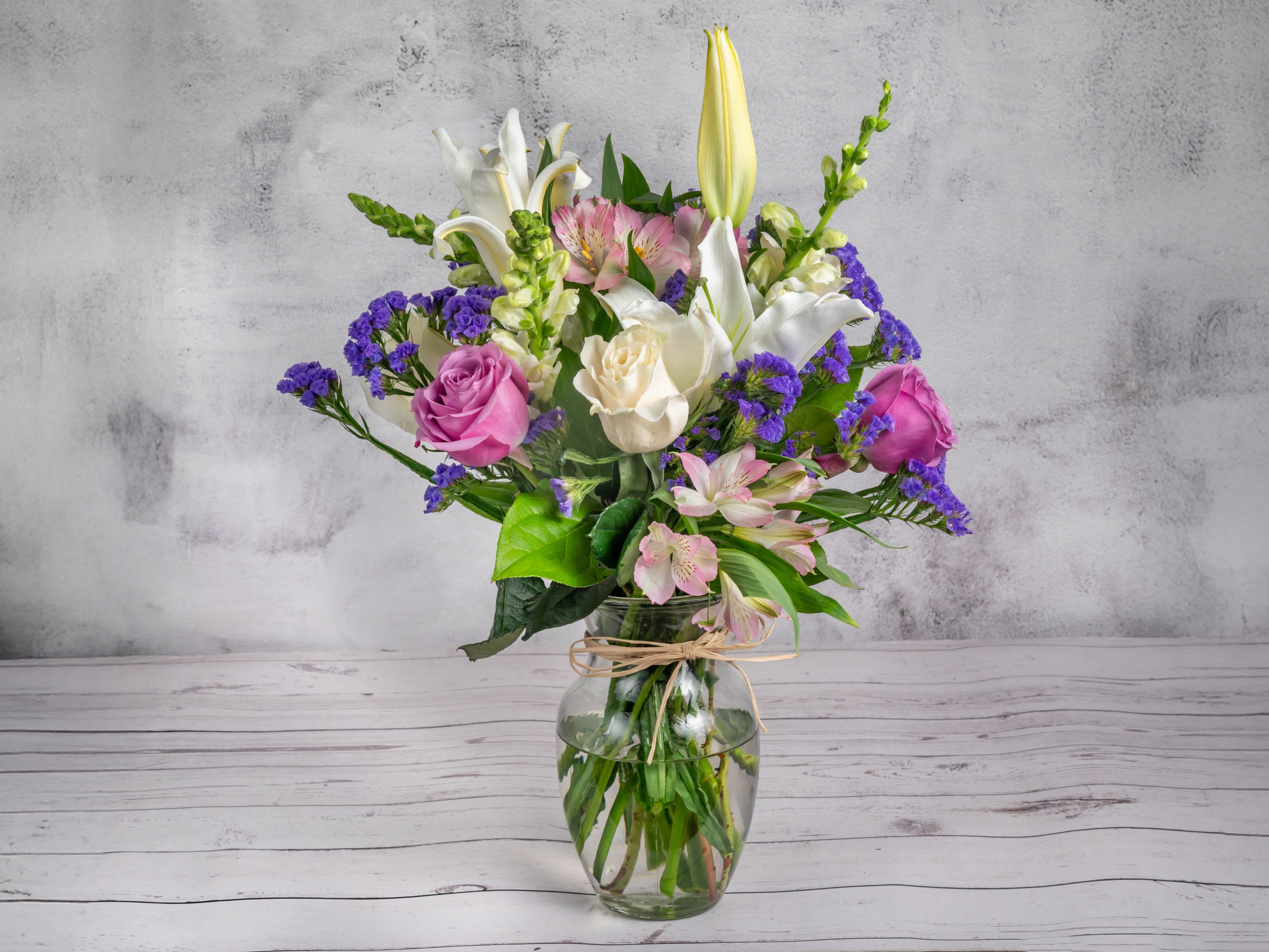 Pastel Whispers Pastel Whispers - A serene blend of white lilies, soft pink alstroemerias, lavender roses, and ivory blooms, highlighted with sprigs of deep purple statice. Arranged in a clear glass vase tied with natural raffia, this bouquet is a perfect symbol of elegance and warmth. Ideal for expressing heartfelt sentiments or adding a touch of grace to any setting.