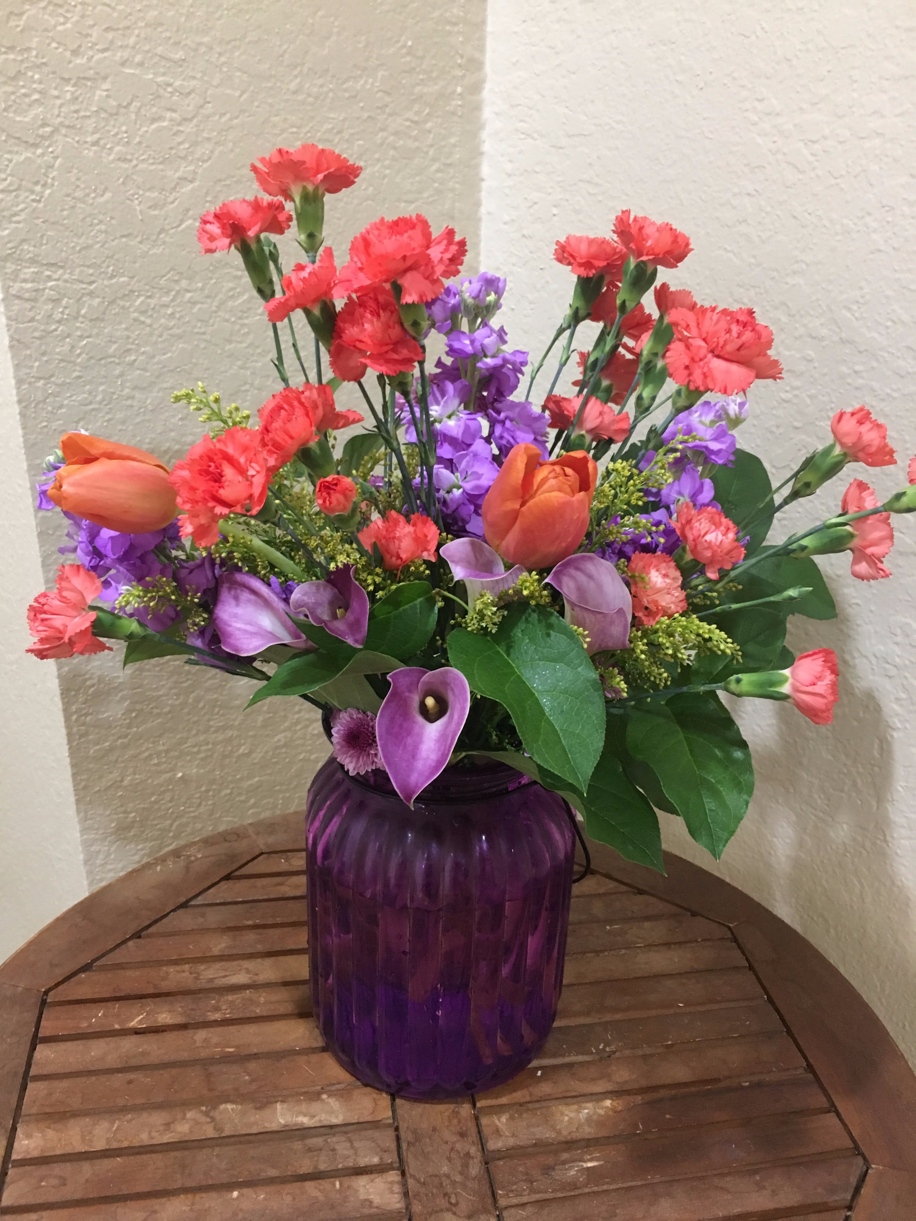 Forest Sunset Forest Sunset - Orange flowers including mini carnations and tulips mixed with purple flowers including purple cala lilies,tulips and fragrant stock in a purple vase.