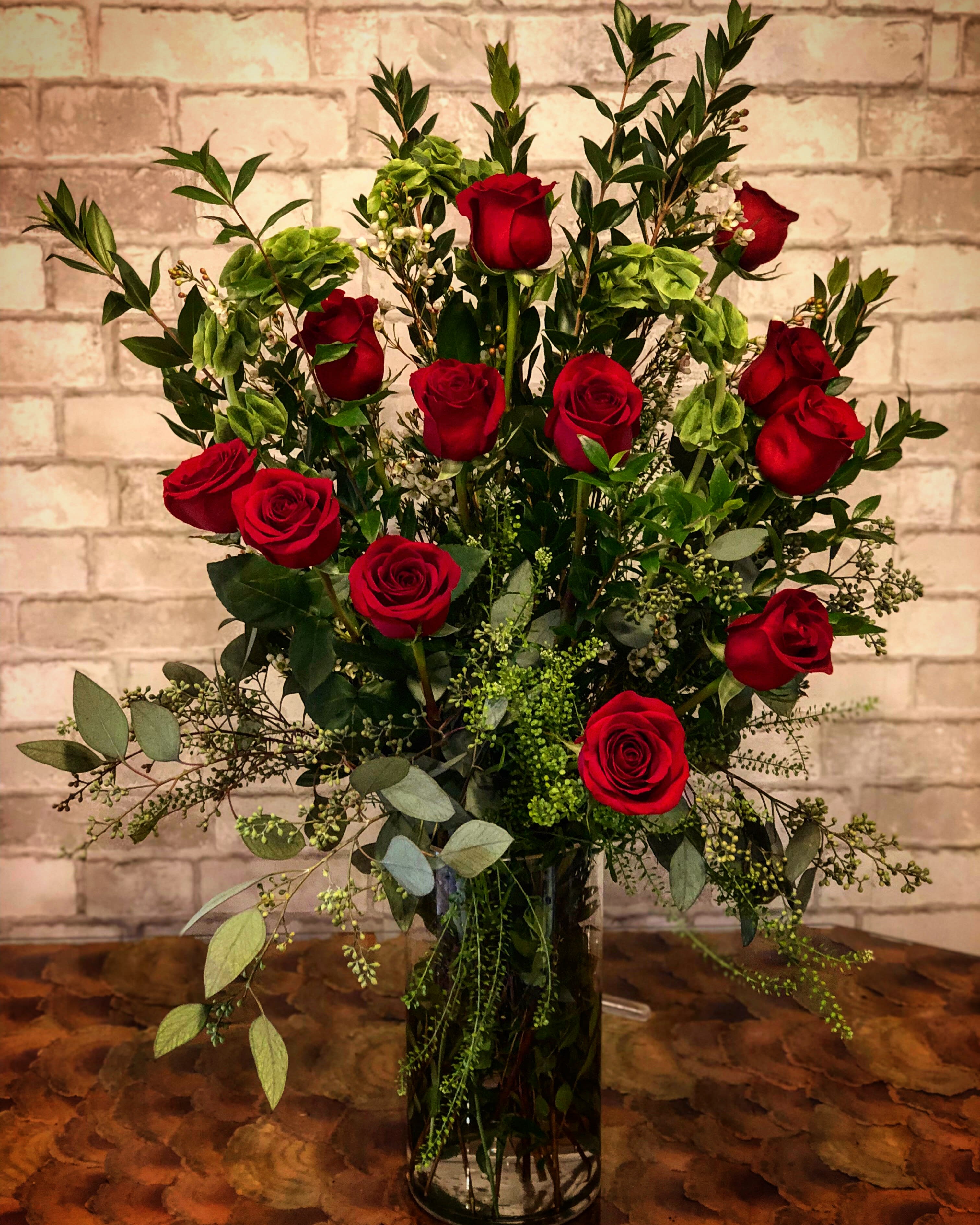 Reds In The Thicket - One dozen long stem red roses arranged in a beautiful bed of lush greenery. Designed in a taller glass vase.
