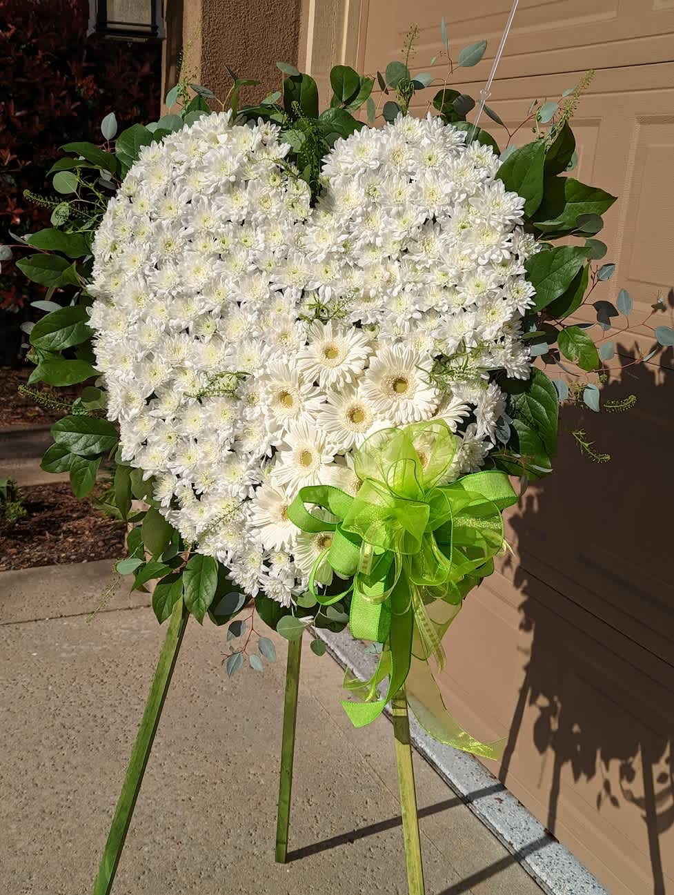 Forever In Our Hearts Standing Spray - Embrace Sympathy with Our 24” Solid Heart Wreath  This 24” solid heart wreath is a poignant tribute, adorned with:  -White mini chrysanthemums -Gerbera daisies -Lemon leaf -Silver dollar eucalyptus  A green ribbon attached at the bottom adds a finishing touch. Ideal for expressing sympathy and honoring the memory of a loved one, this elegant arrangement conveys comfort and support during difficult times.