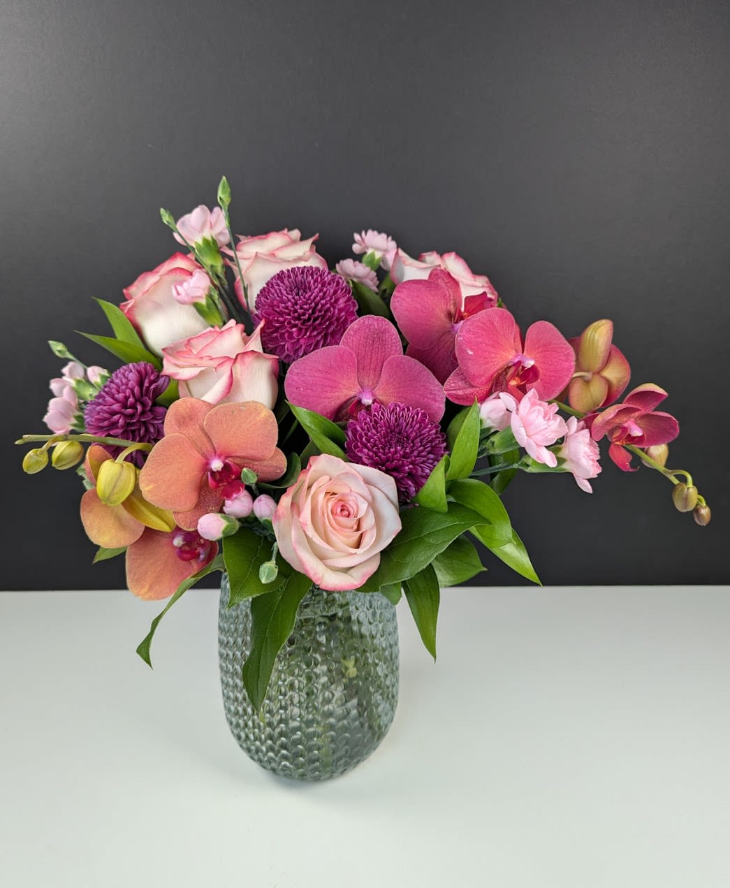 Lavish Orchid Dusk - A striking display of luxury and warmth, Lavish Orchid Dusk captures the ethereal beauty of a sunset with its rich, layered hues. This opulent arrangement is designed to evoke elegance and sophistication, making it a statement piece for any occasion Featuring:  -Sunset Phalaenopsis orchids  -Blush roses  -Purple mums  -Light pink mini carnations  Soft, romantic tones blend seamlessly with deep, regal shades, creating a captivating composition that radiates refinement. Perfect for those who appreciate timeless beauty and exquisite floral artistry.         