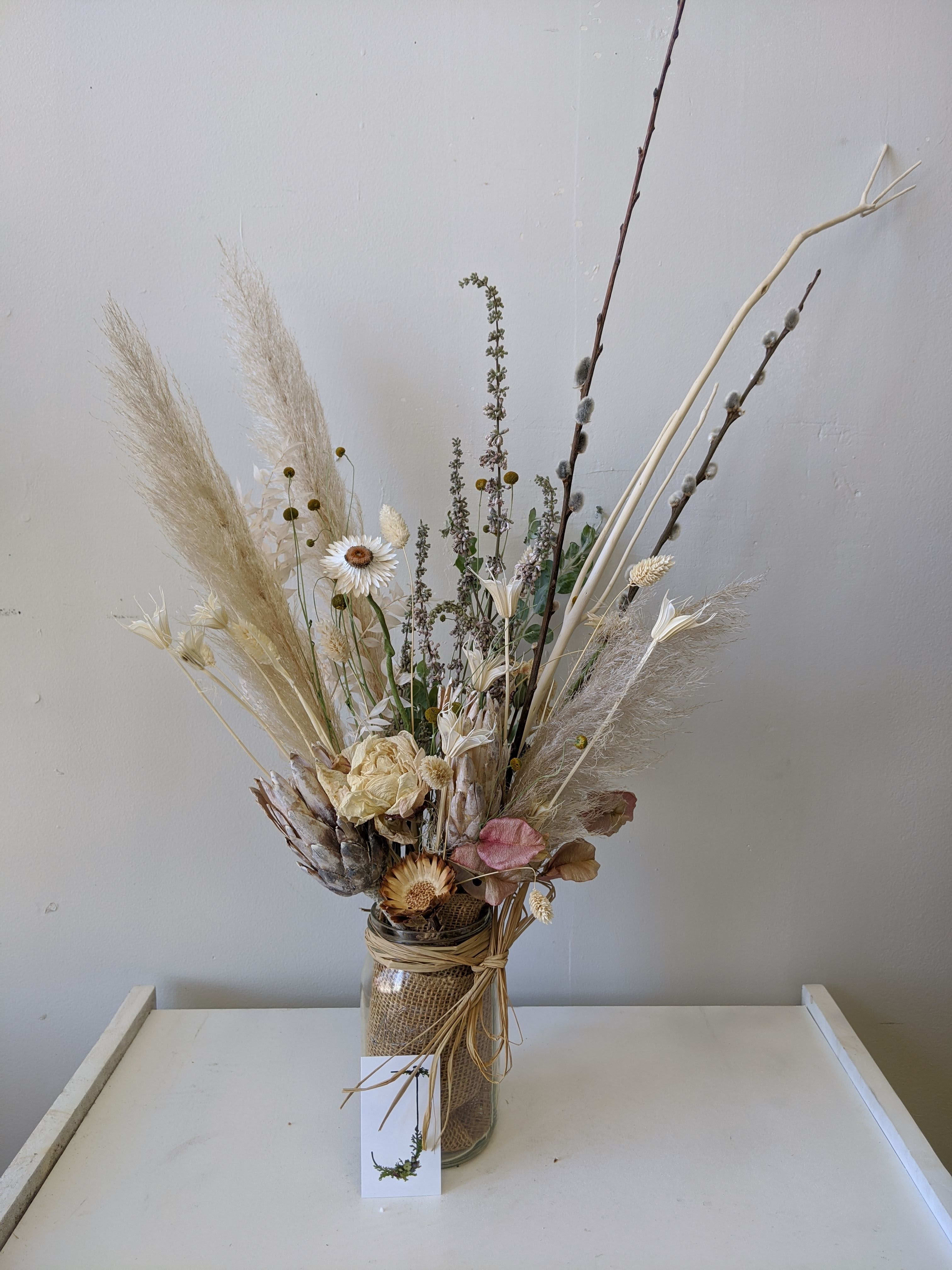 Everlasting Mason Jar - This everlasting dried arrangement includes dried Peonies and pampas and comes in a mason jar. Business card in photo for scale. Note that because of the nature of dried flowers, some may break and crumble if not carefully handled.