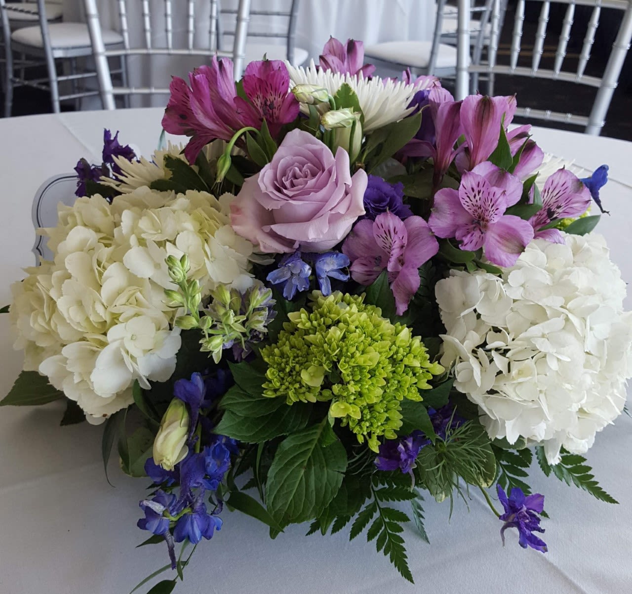 Lavender Kiss - Beautiful White and Green Hydrangea Nestled in a Bed of Lush Foliage and Accented With Blue Delphinium, White Fuji Mums, Purple Roses and a “kiss” of Purple Alstroemeria. This is a Stunning Bouquet for the Home, on the Dinner Table or That Special Event as a Centerpiece for Wedding or Shower.