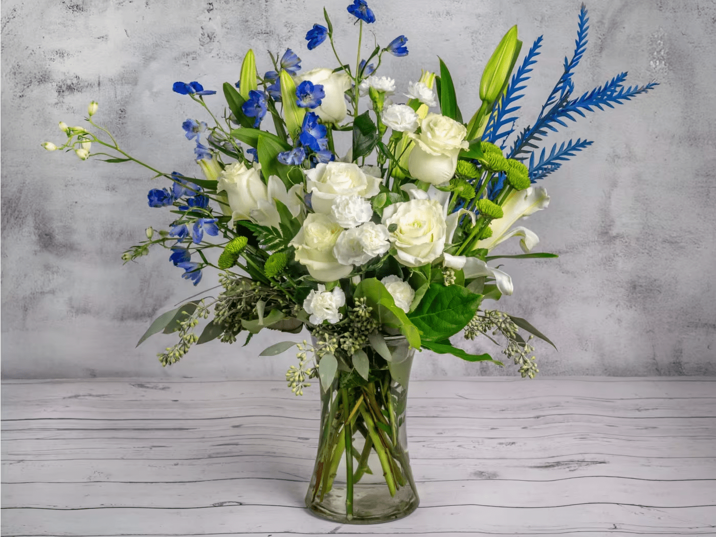Heavenly Breeze - Elevate any space with this serene and elegant flower arrangement. Featuring pristine white roses, lilies, and carnations, accented with delicate blue delphiniums and lush greenery, this bouquet captures the essence of tranquility and sophistication. Arranged in a sleek clear glass vase, this design offers timeless beauty, perfect for expressing sympathy, celebrating milestones, or adding a touch of grace to your home. The soft whites and vibrant blues blend harmoniously to create a striking yet calming effect.
