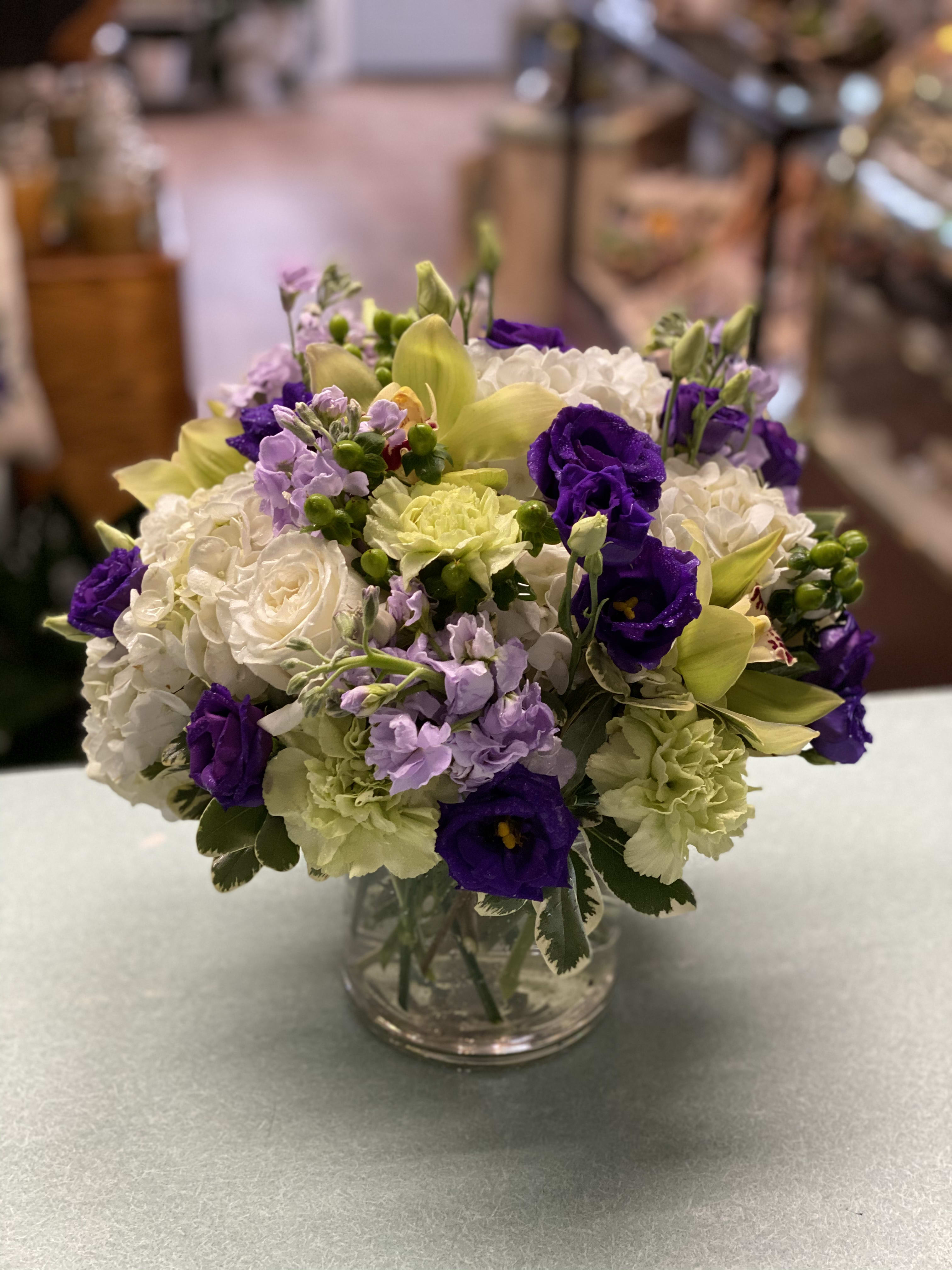 Elegant Spring - Green Cymbidium Orchids, White roses, White hydrangea, purple lisianthus, lavender stock, green carnations, green hypericum berries, in a clear glass cylinder
