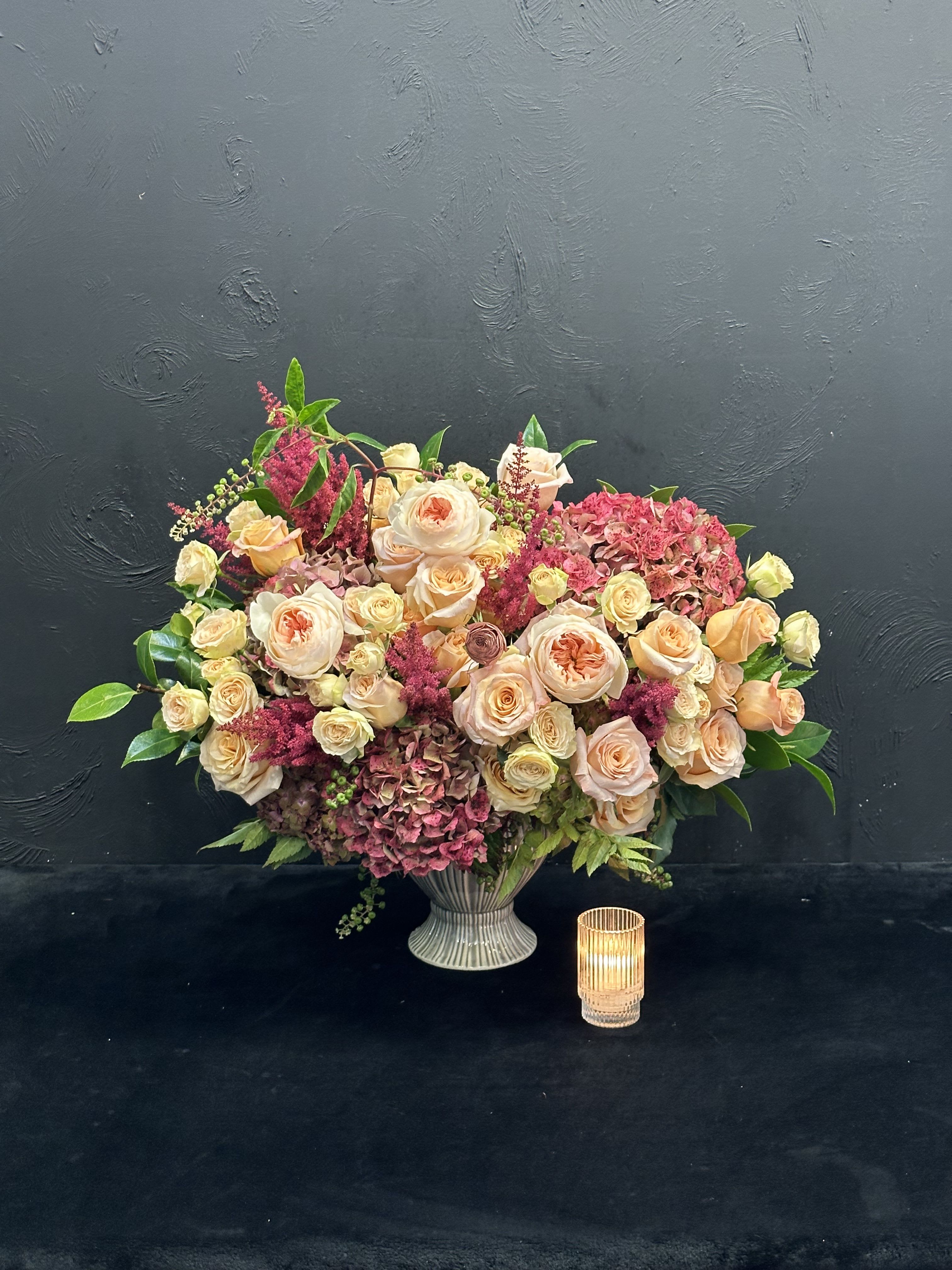 Urn of Delights  - This stunning arrangement showcases peach garden roses, lush hydrangeas, and delicate spray roses, all beautifully nestled in an elegant urn. The garden roses, with their velvety petals, exude a soft charm, while the hydrangeas provide a generous, textured backdrop in creamy white and soft peach tones. Interspersed among them, the spray roses add playful accents, enhancing the overall romance of the composition. The combination of these flowers creates a warm and inviting display, perfect for adding a touch of sophistication to any space or occasion.    