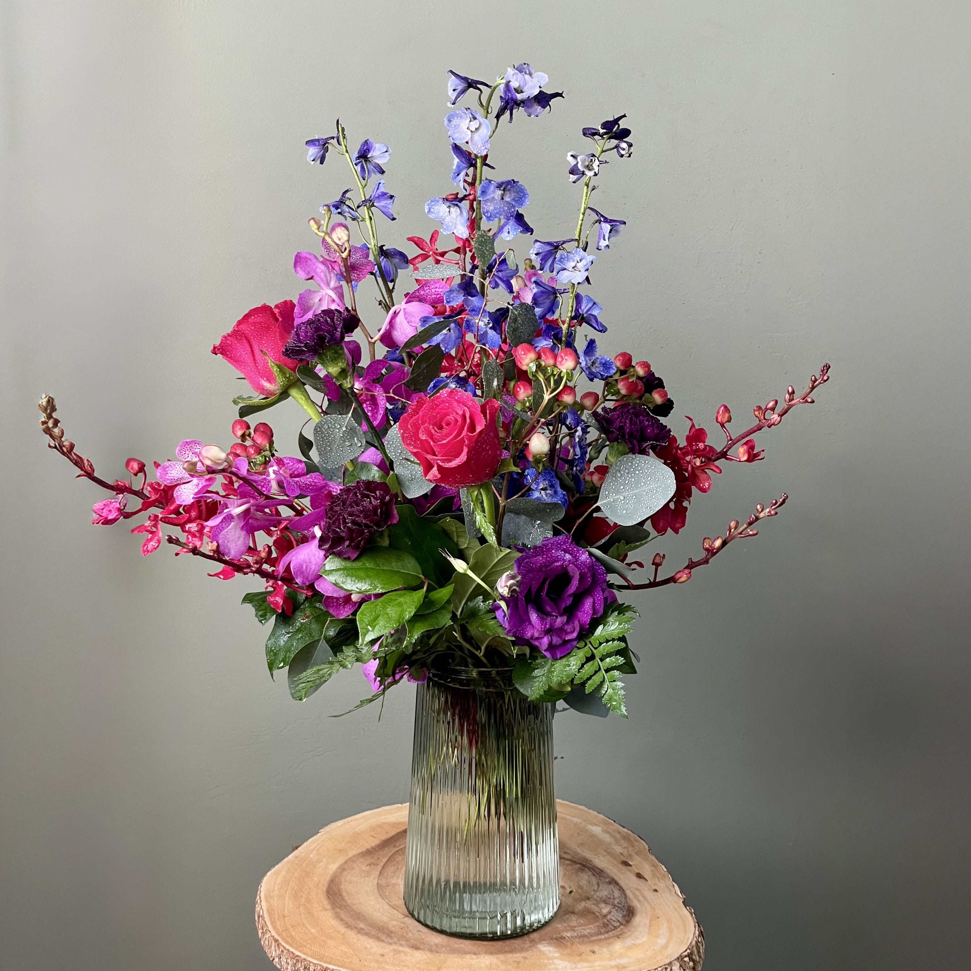 Falling For You (SF551) - Captivate their heart with this enchanting arrangement of deep purple and fuchsia tones. Featuring dark blue delphinium, hot pink roses, Moonvista carnations, purple mokara orchids, James Storie orchids, red hypericum berries, and purple lisianthus. all elegantly arranged in a clear ribbed glass vase. A breathtaking display of romance and charm!