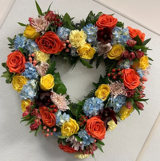 Uplifting Love - This colorful and bright heart wreath spray is representative of a life lived to its fullest. Colors and Flowers may change based on season and availability.