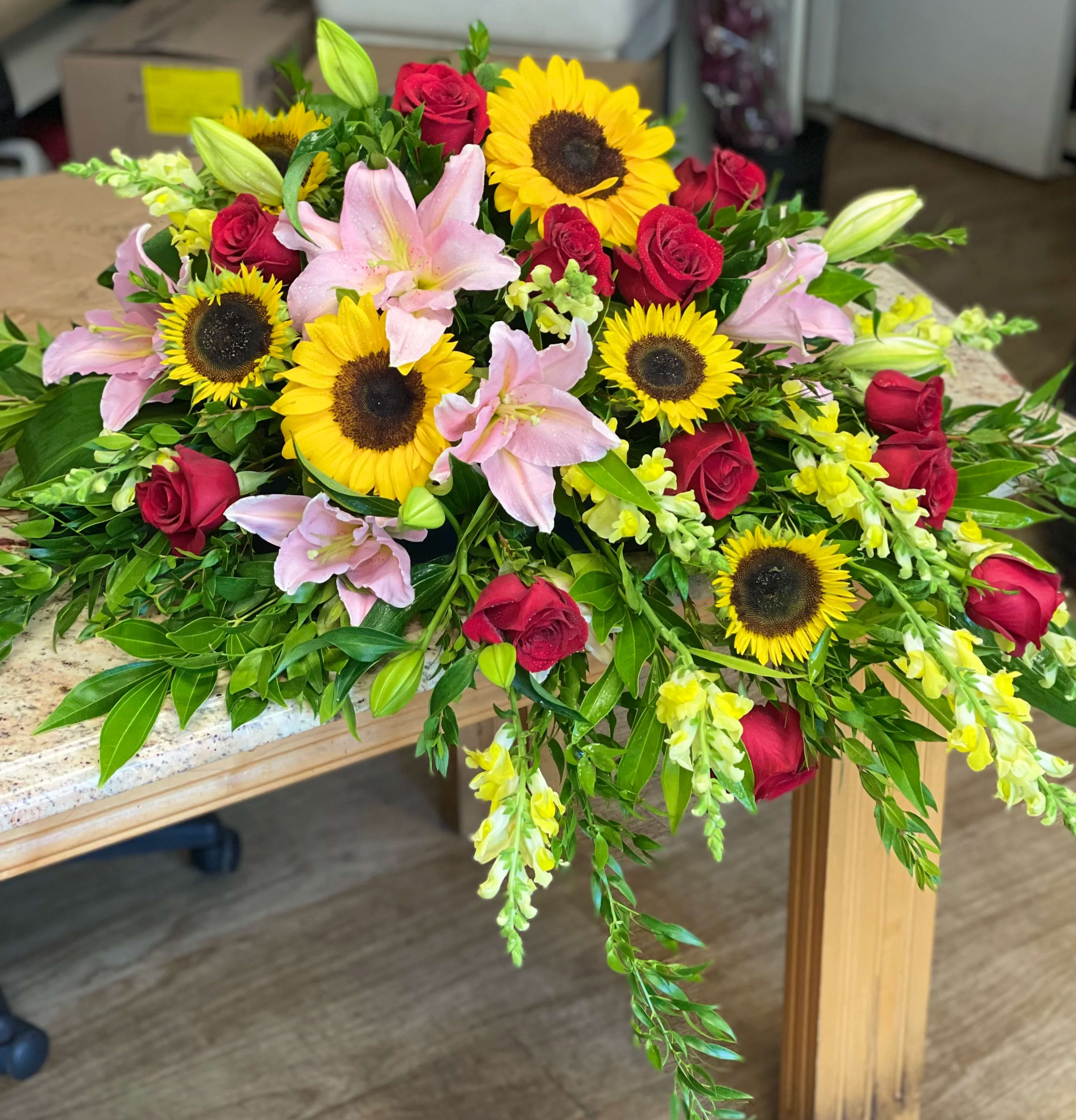 Sunflower Love Casket Spray - Foliage: Sunflower, Rose, Lilly, Snapdragon and mix emerald greenery   We advise our customers to order 24-48 hours in advance of their scheduled delivery to ensure that we are able to have similar flowers and colors in the shop for their order.  $350 (standard) as shown