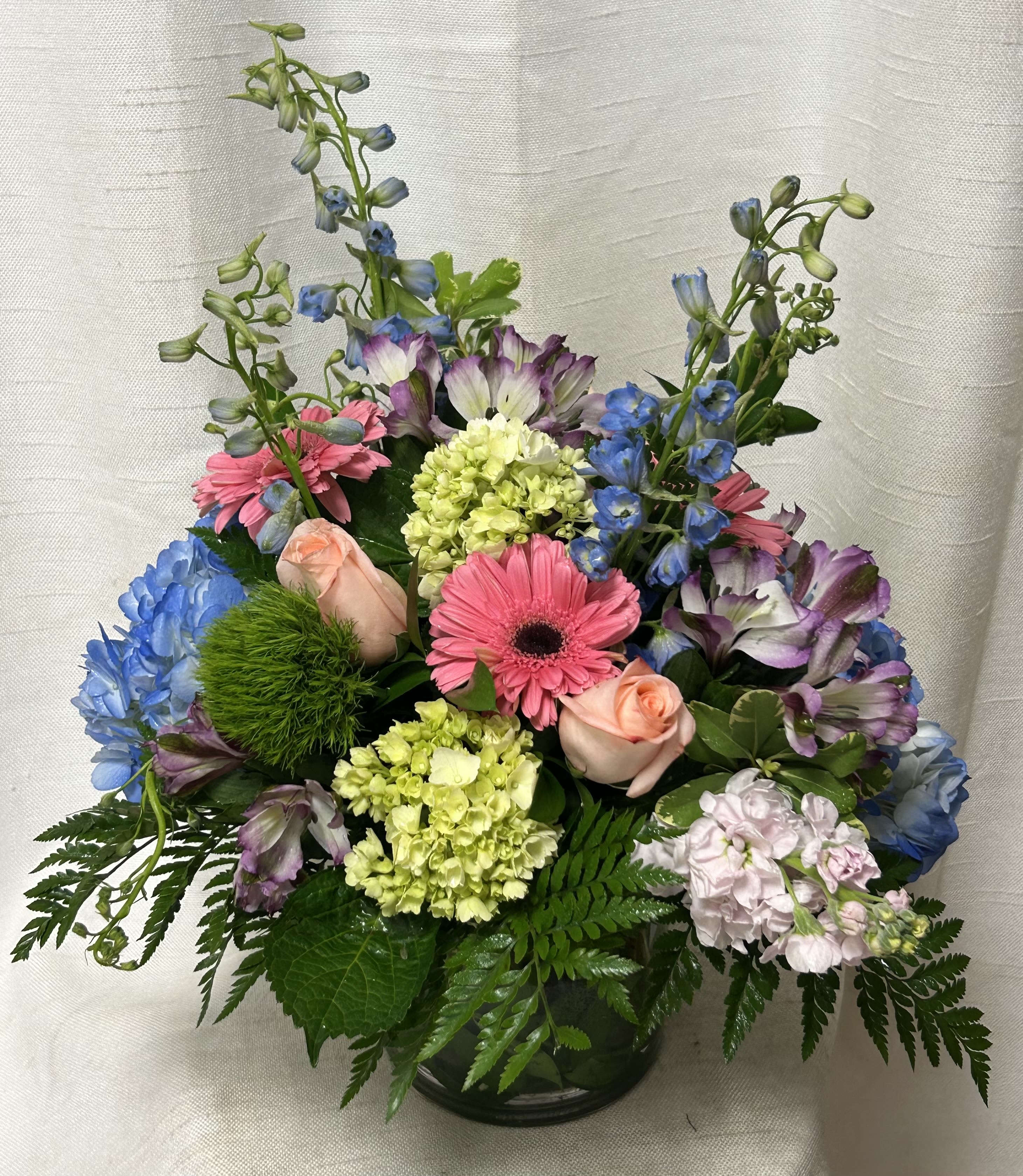 Serenity - Lush arrangement is created in a 6x6&quot; glass cylinder vase. We combine blue and green Hydrangea, Gerberas, Roses, Delphinium, mini Calla Lilies and more. Comes professionally designed by our talented designers