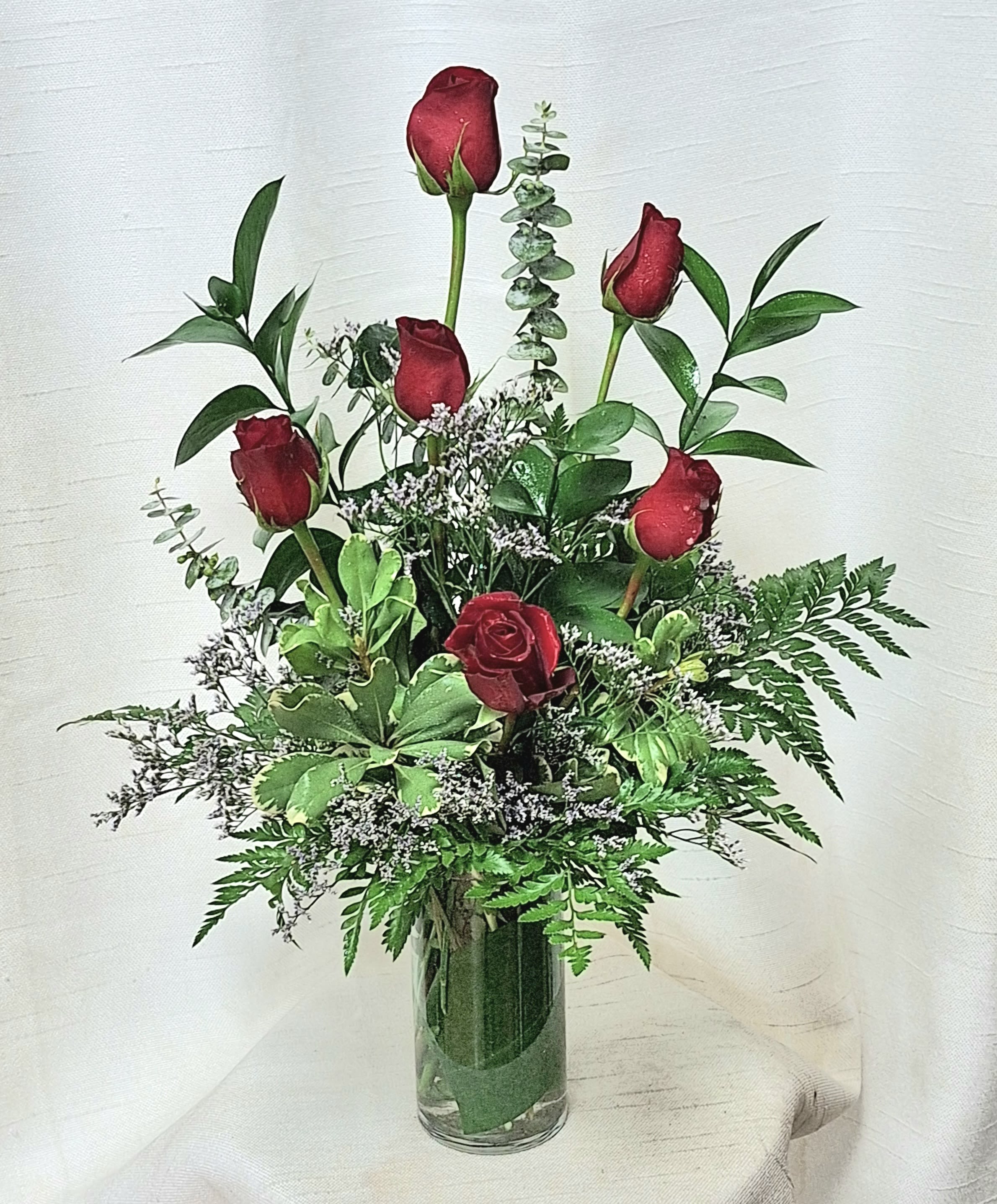 Elegance - Revel in the allure of our modern floral arrangement- featuring six stunning red roses arranged in a leaf-lined glass cylinder vase, accentuated with assorted greenery. This captivating display is available in your choice of red, white, pink, lavender, or yellow, offering versatility to suit any personal style or occasion. Add an instant uplift to any space with this perfectly balanced and charming arrangement. 