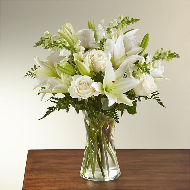 Eternal Rest - Introducing the &quot;Eternal Rest&quot; arrangement, a serene and heartfelt tribute to honor the memory of a loved one. This arrangement features a delicate combination of white roses, white lilies, white snapdragons, accompanied by lush greens and filler, all thoughtfully arranged in a clear glass vase.  