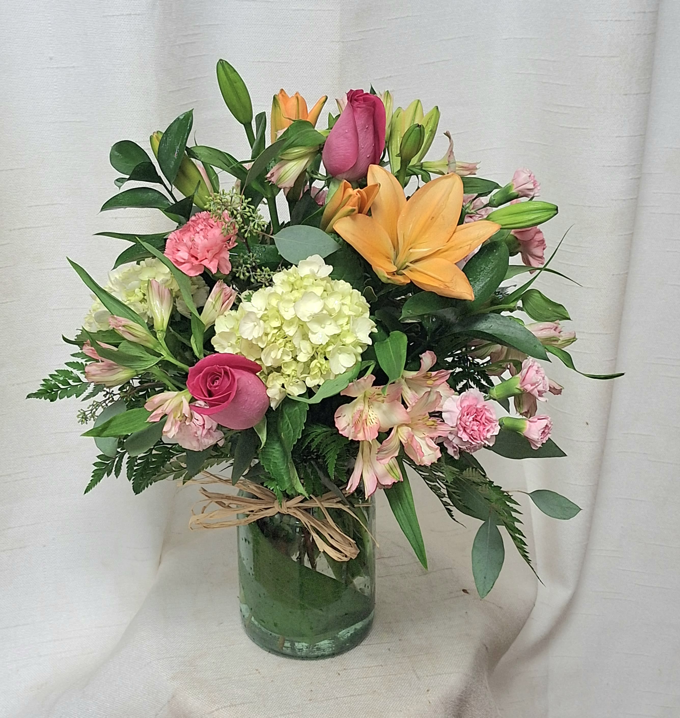 Life is Good - Introducing &quot;Life is Good,&quot; a delightful flower arrangement that combines vibrant shades of orange and pink to create a harmonious and stunning display. This arrangement features a captivating blend of orange lilies, pink roses, mini pink spray roses, pink alstroemeria, mini green hydrangea, lush greenery, and filler flowers, all artistically arranged in a traditional style vase.  