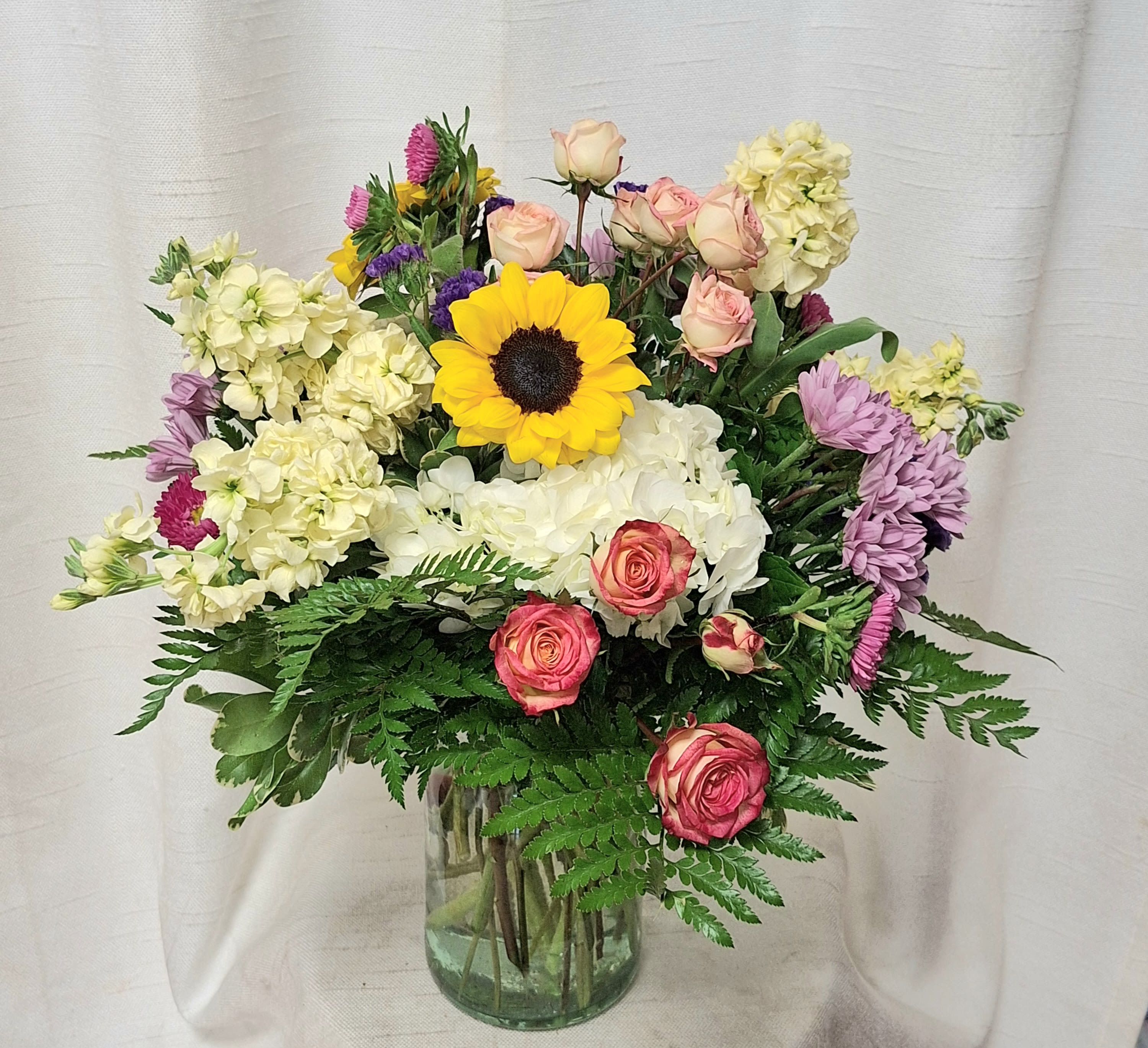 Colorful Surprise - Experience a burst of vibrant hues with our Colorful Surprise arrangement. This stunning ensemble features an array of flowers including Stock, Sunflowers, Spray Roses, Hydrangea, Motsumoto Asters, and assorted greenery, all artfully arranged in a clear glass vase. Perfect for adding a pop of color and joy to any space, this arrangement is sure to delight and impress. 
