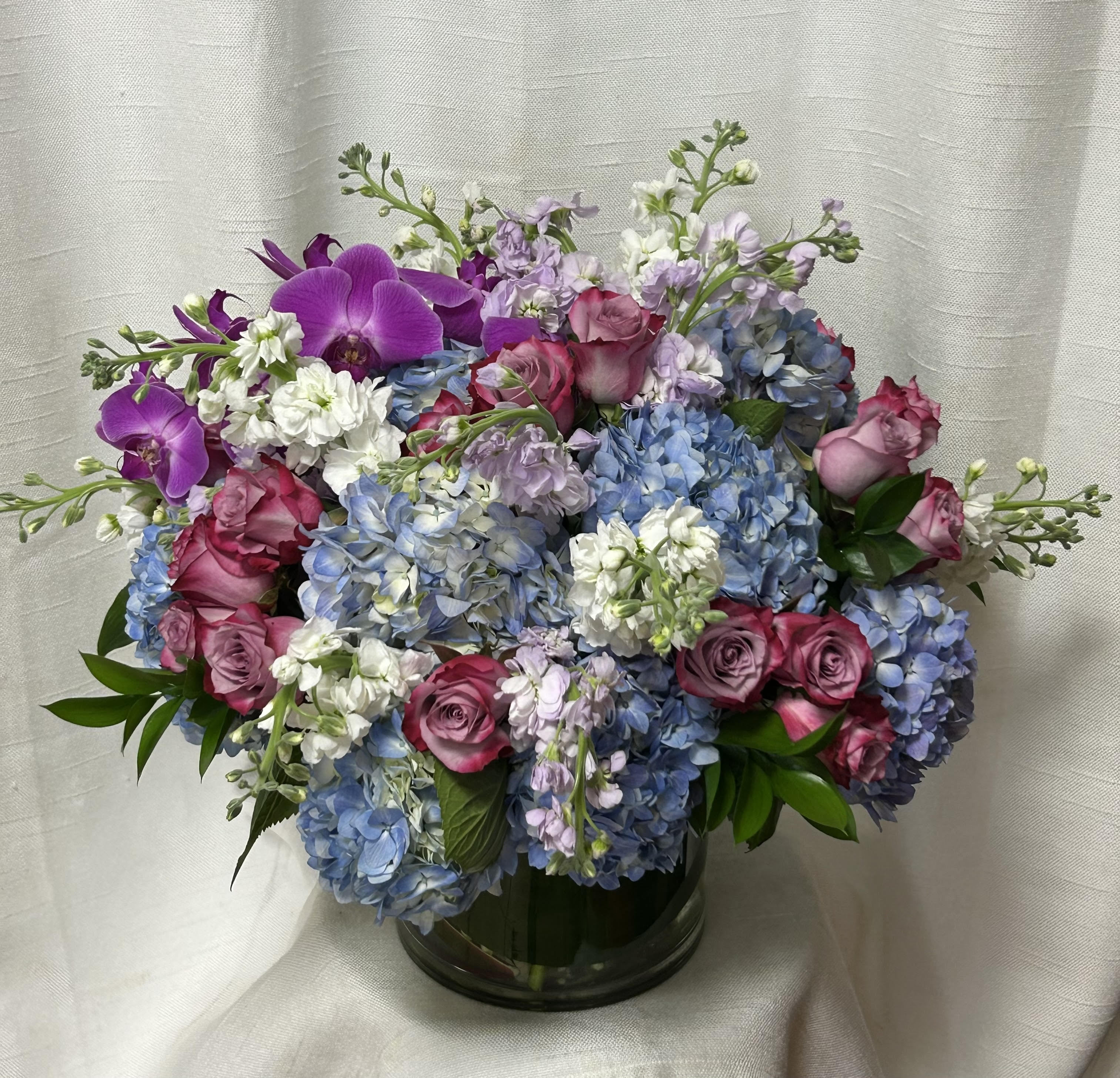 Grand Prize (BB) - Presenting an opulent display designed to make a lasting impression. This luxurious arrangement features a sophisticated blend of blue hydrangea, lavender roses, purple and white stock, lush greens, and filler flowers, all artistically arranged in an 8x8 glass vase.