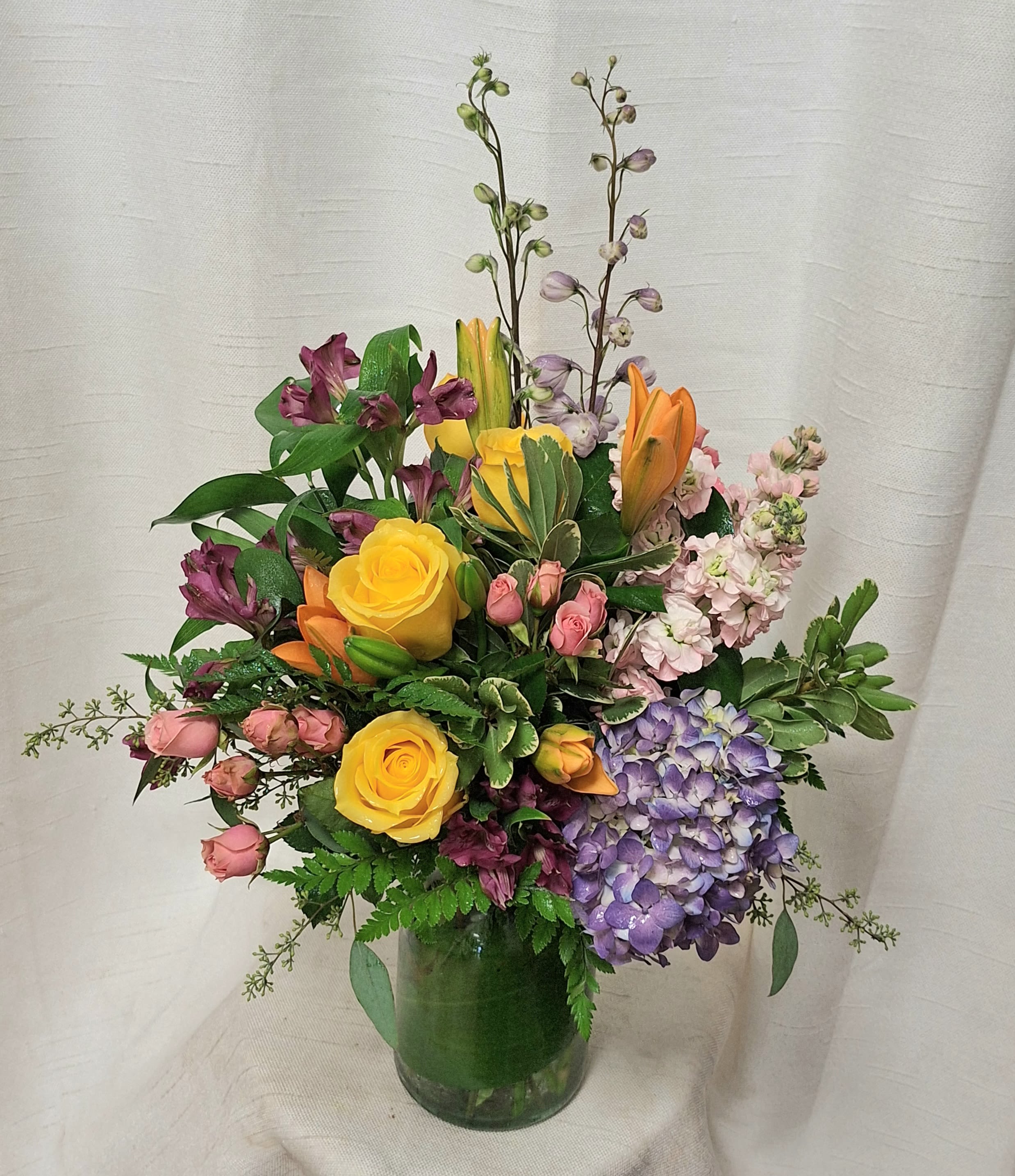 Happy Days (BB) Happy Days (BB) - Elevate your space with our modern floral arrangement, thoughtfully crafted in a clear glass leaf-lined vase. This artistic ensemble features a vibrant mix of orange lilies, delphinium, stock, spray roses, purple hydrangea, yellow roses, and assorted greenery. Captivating in its design, this arrangement brings a burst of color and elegance to any room, making it a perfect statement piece for any occasion.