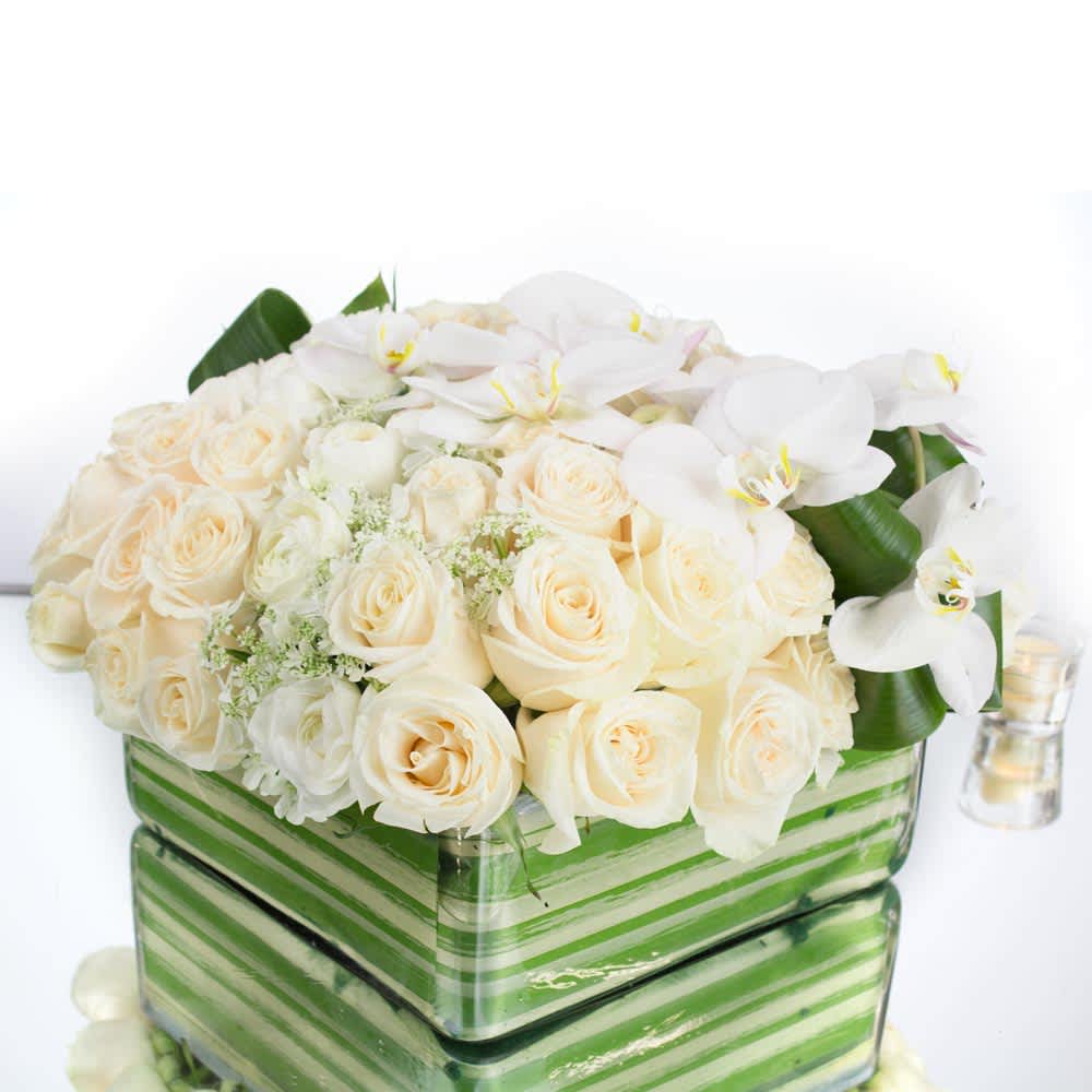 Pure White (BB) - During this time of mourning, our funeral flower arrangement offers a heartfelt tribute that honors your loved one's memory with grace and beauty. This exquisite arrangement includes a selection of white roses, white Phalaenopsis blooms, white ranunculus, and filler greens, expertly arranged in a square glass vase. To add additional accents and details, we incorporate thoughtful elements that enhance the overall aesthetic and symbolism of the arrangement.