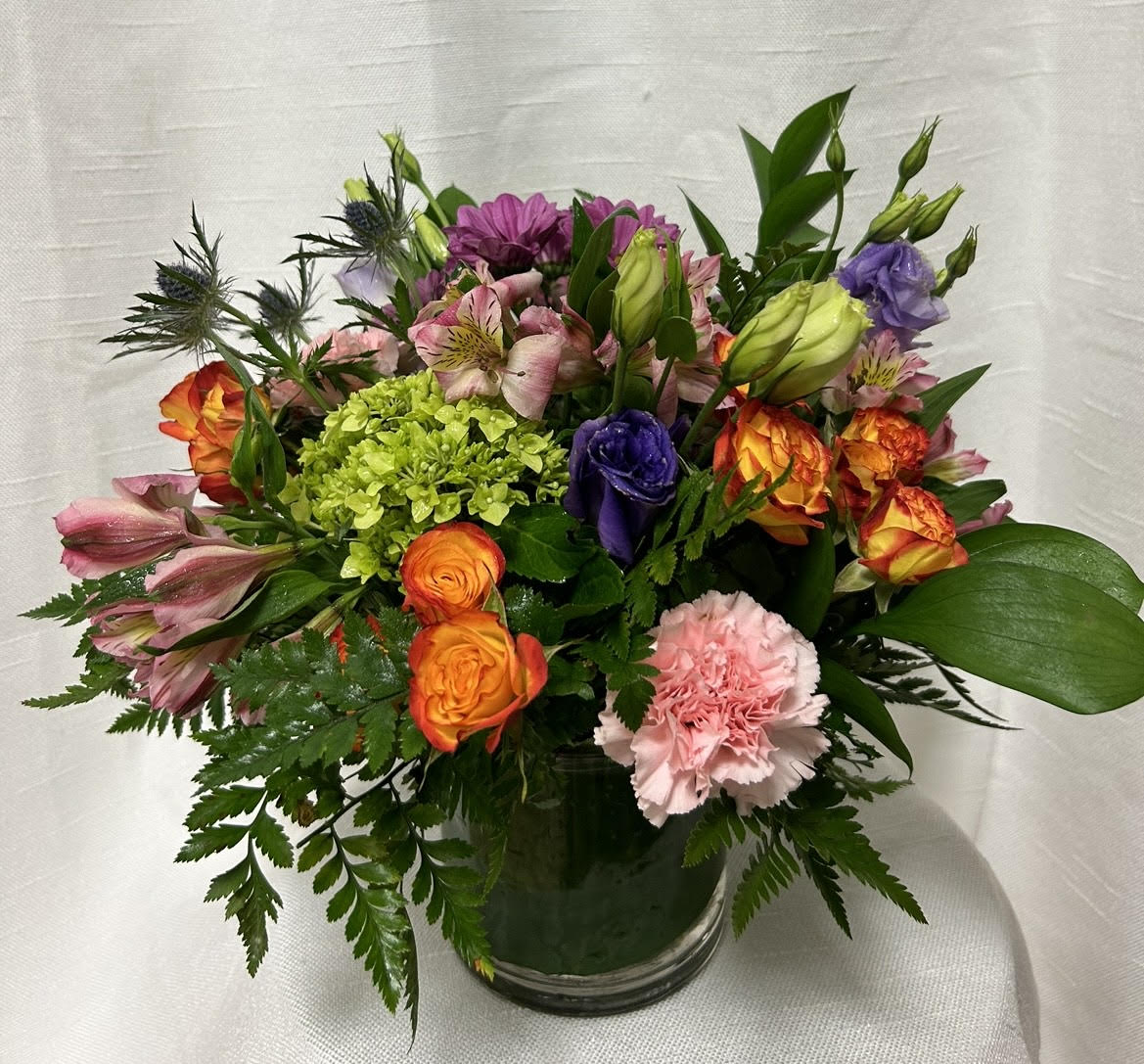 Party Punch (BB) - Introducing a modern and chic floral arrangement that exudes contemporary elegance. This arrangement showcases a unique combination of green mini hydrangea, orange spray roses, blue thistle, pink carnations, pink alstroemeria, and hot pink gerber daisies, all expertly arranged in a leaf-lined glass cylinder vase.